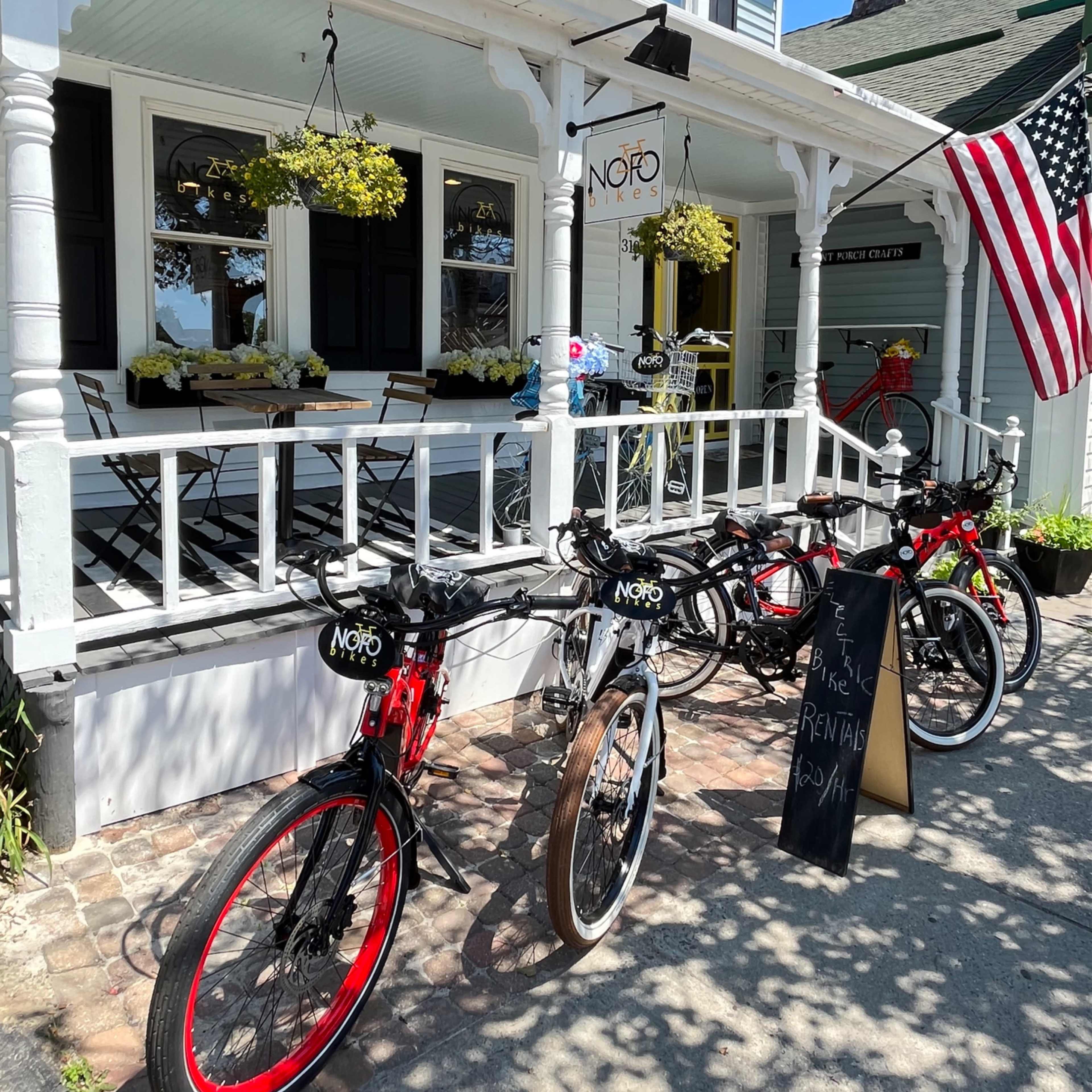 A row of bicycles is parked in front of a building with a sign that reads "NOFO" and hanging flower planters, alongside an American flag.