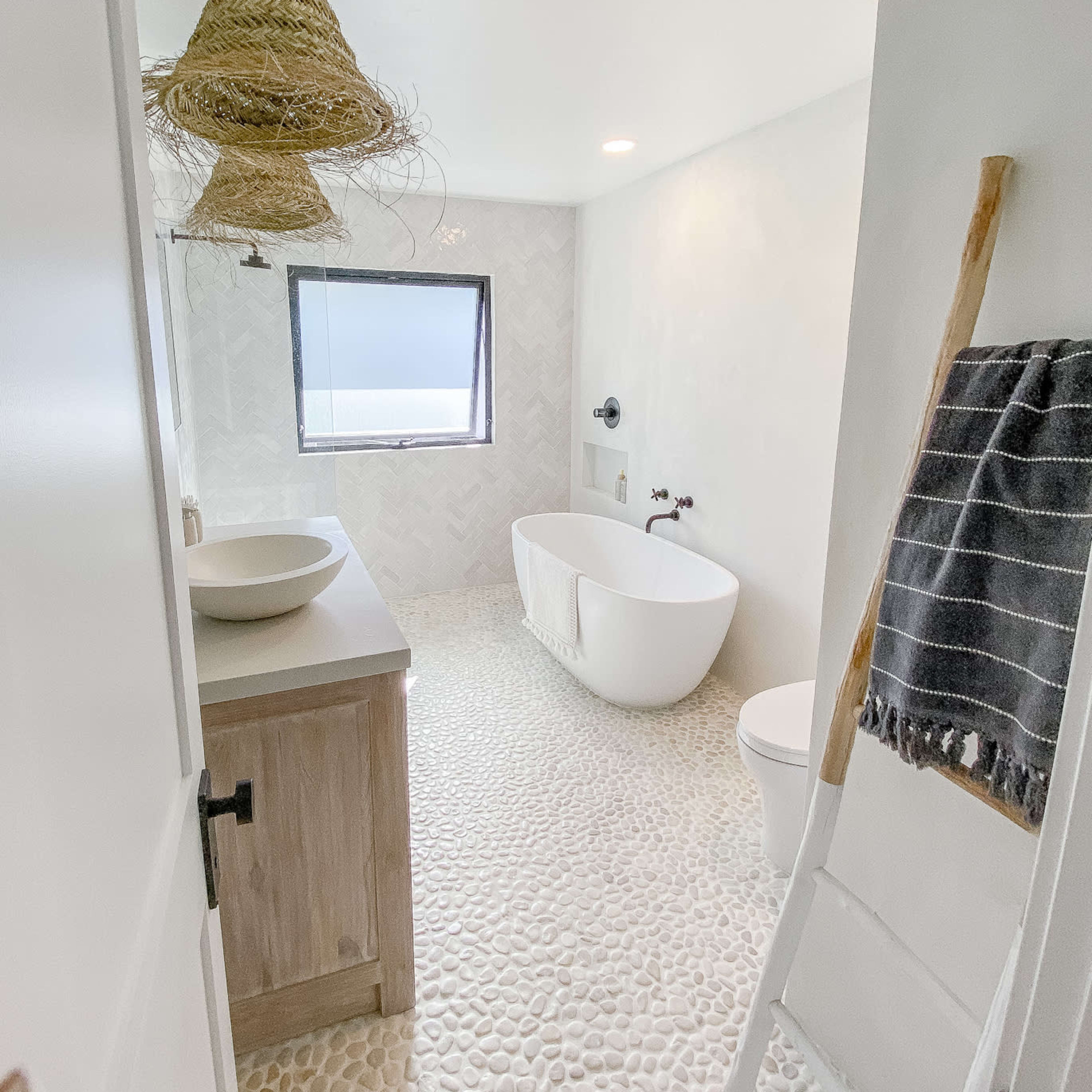 A modern bathroom with a freestanding bathtub, a wooden vanity, a pebble stone floor, and a large window.