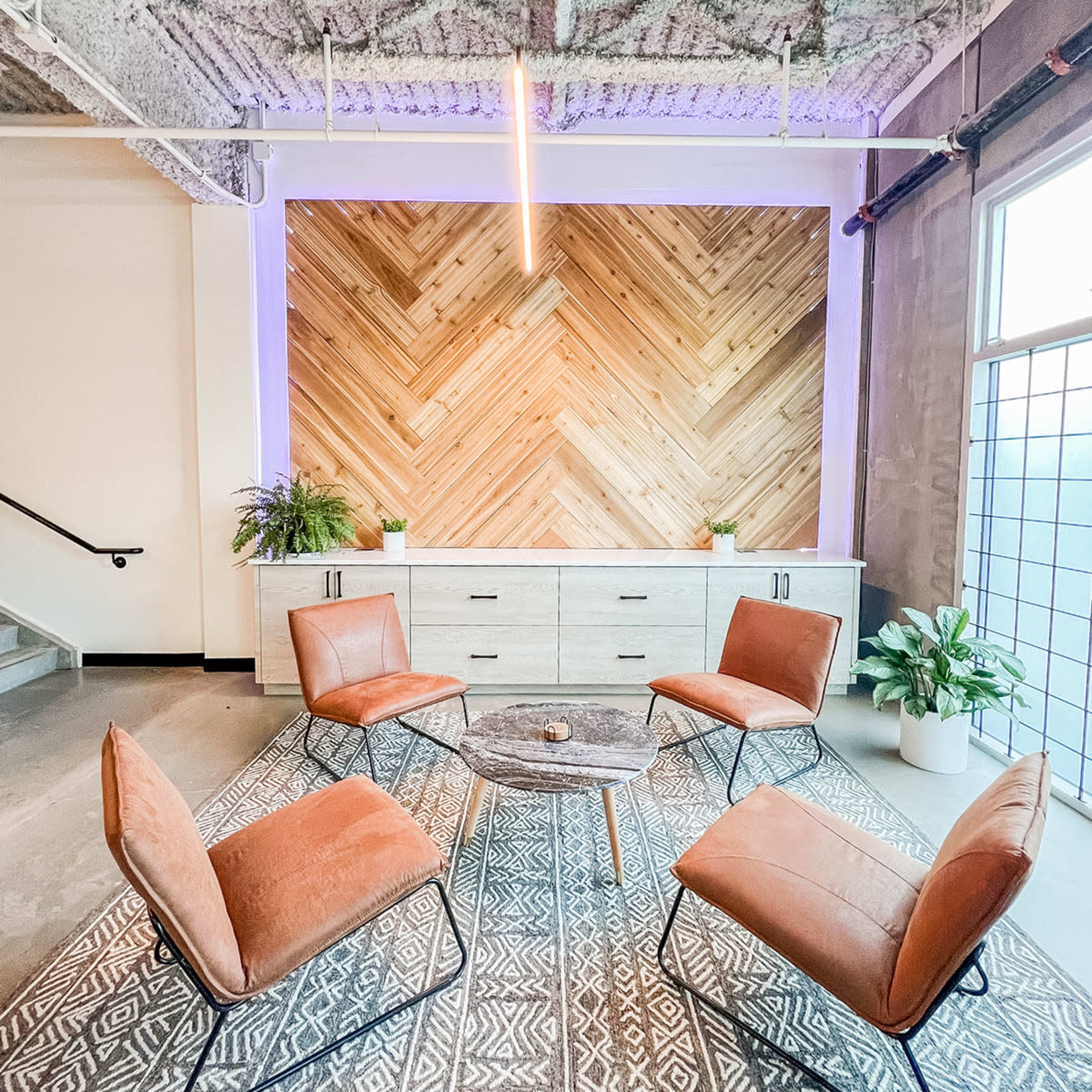 A modern lounge area featuring four rust-colored chairs arranged around a circular wooden table, set against a backdrop of a wooden accent wall with a chevron pattern.