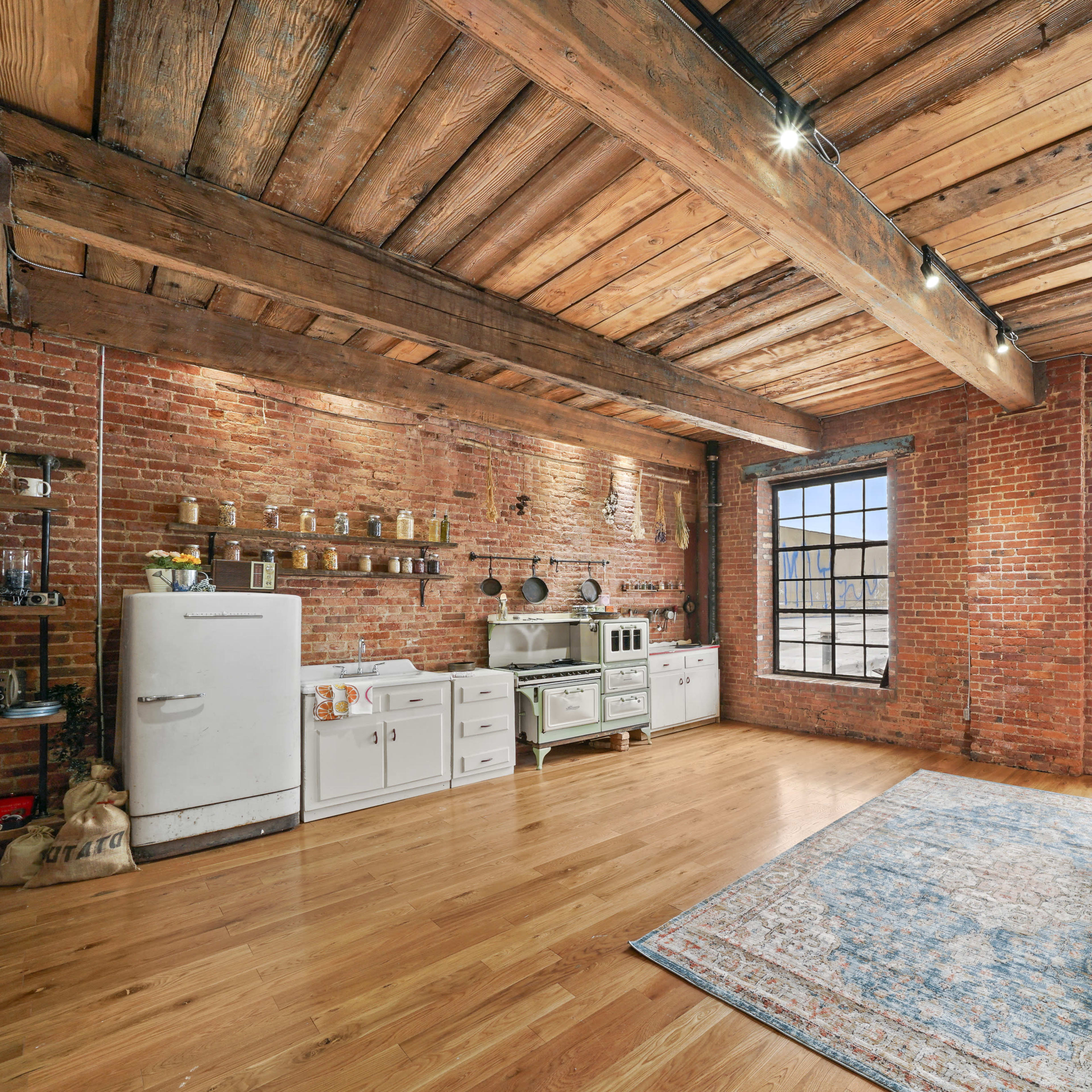 A spacious interior with exposed brick walls, wooden beams, and large windows, furnished with a vintage couch and a retro kitchen setup.