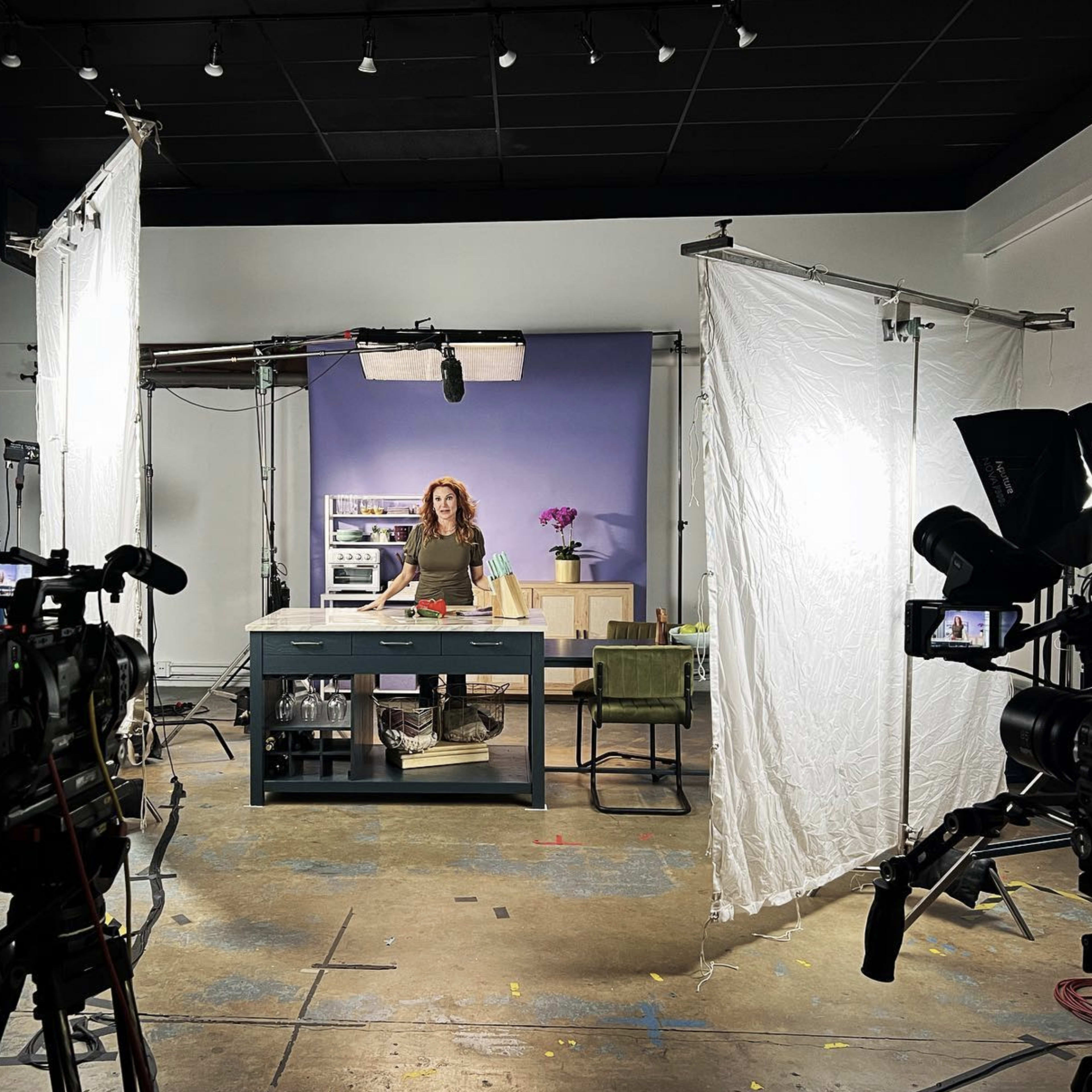 A video production set features a woman standing behind a table surrounded by cameras, lights, and backdrop elements.