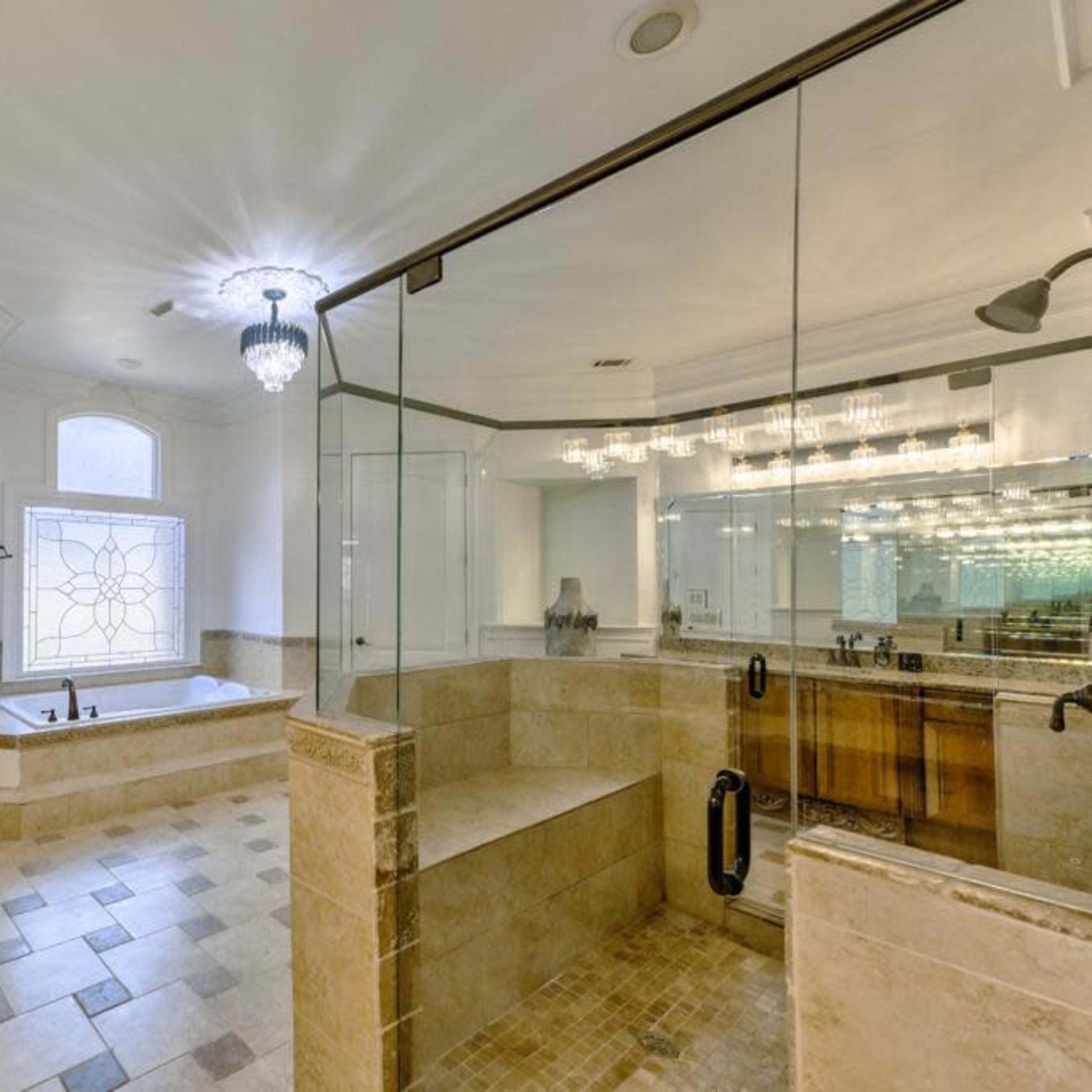 A spacious bathroom featuring a glass-enclosed shower, a large soaking tub by a window, and a double vanity with multiple light fixtures.