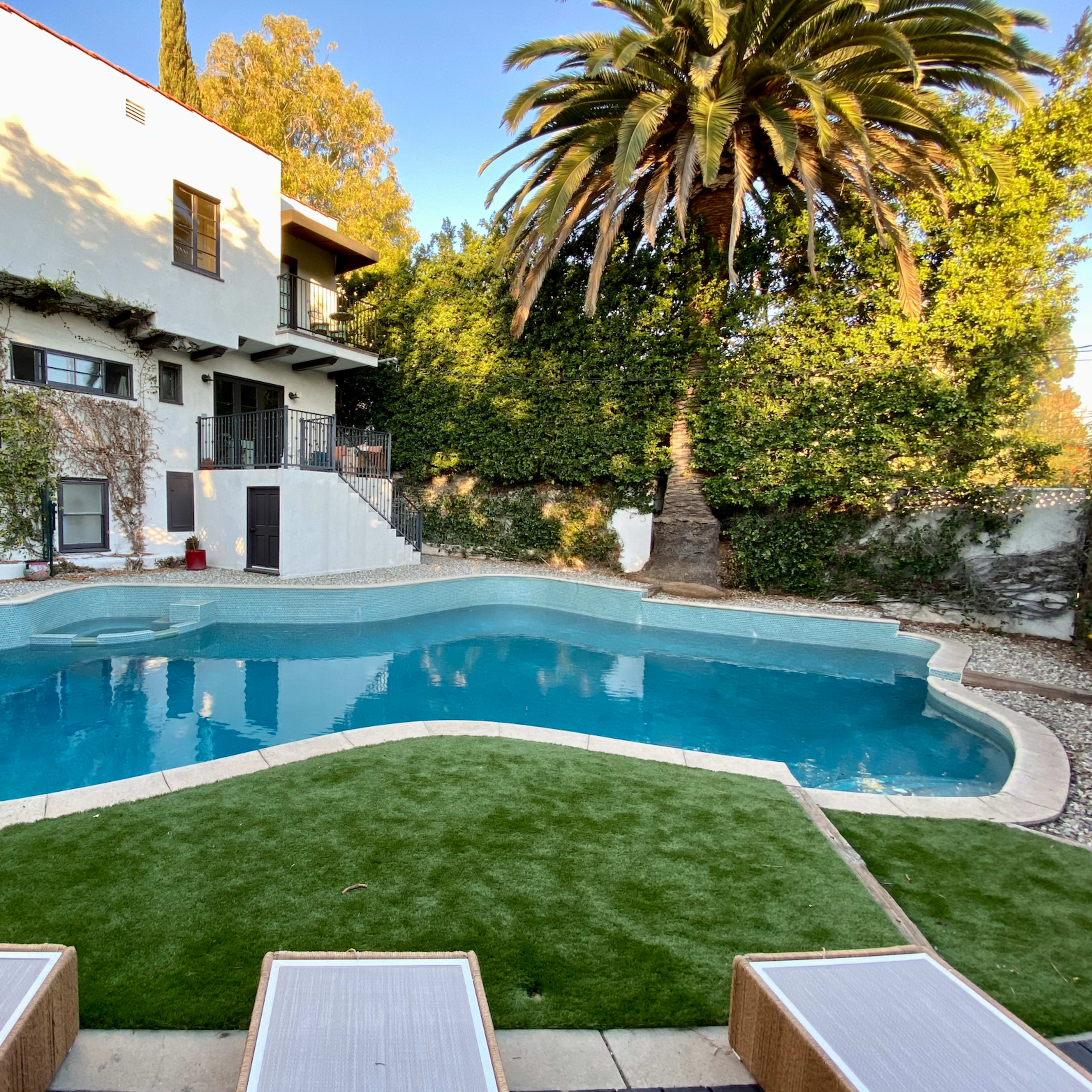 A sparkling blue swimming pool surrounded by a landscaped area featuring palm trees and a multi-level house in the background.