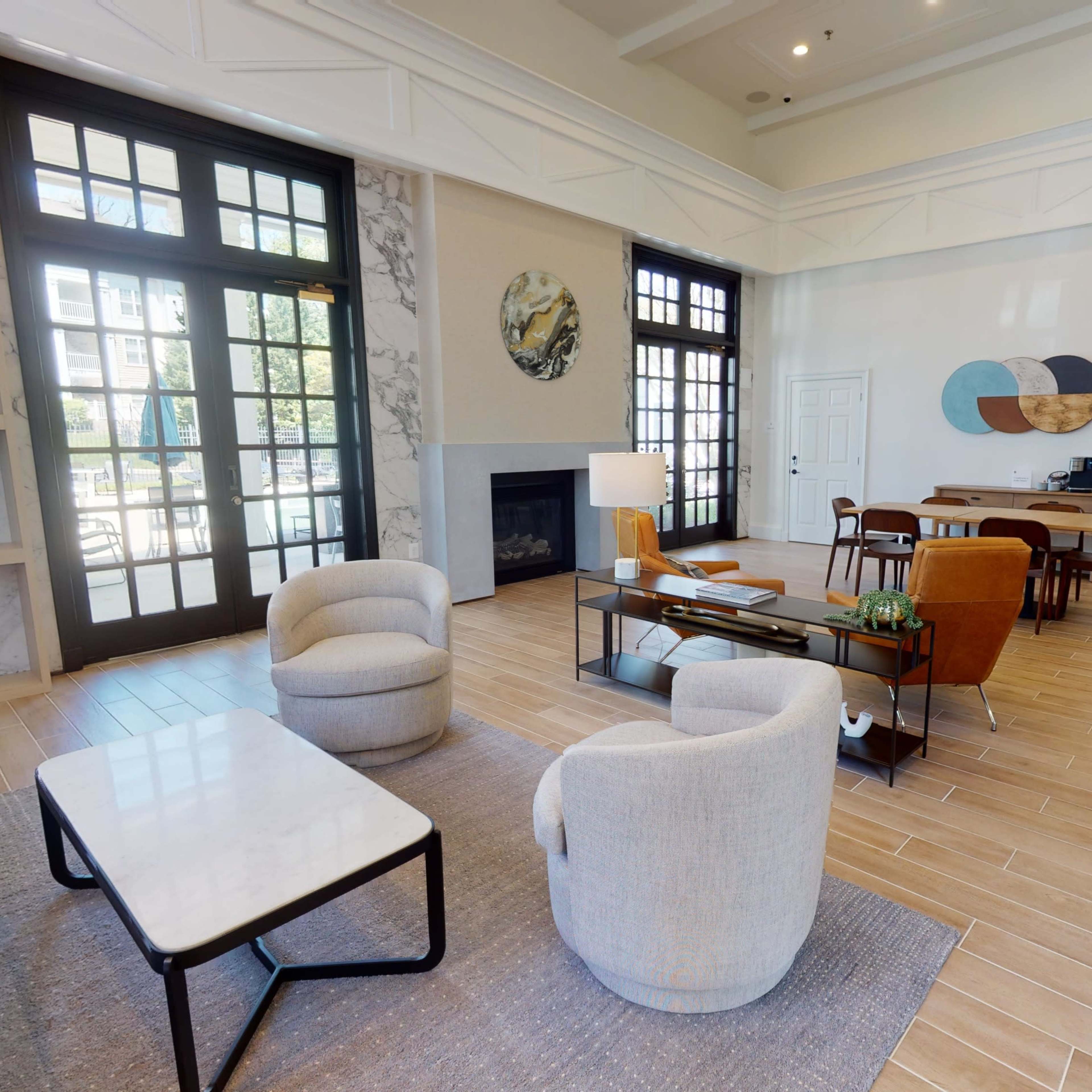 A spacious, modern lounge area featuring soft seating, a coffee table, and a dining space with wooden chairs and a table, surrounded by large windows and decorative elements on the walls.