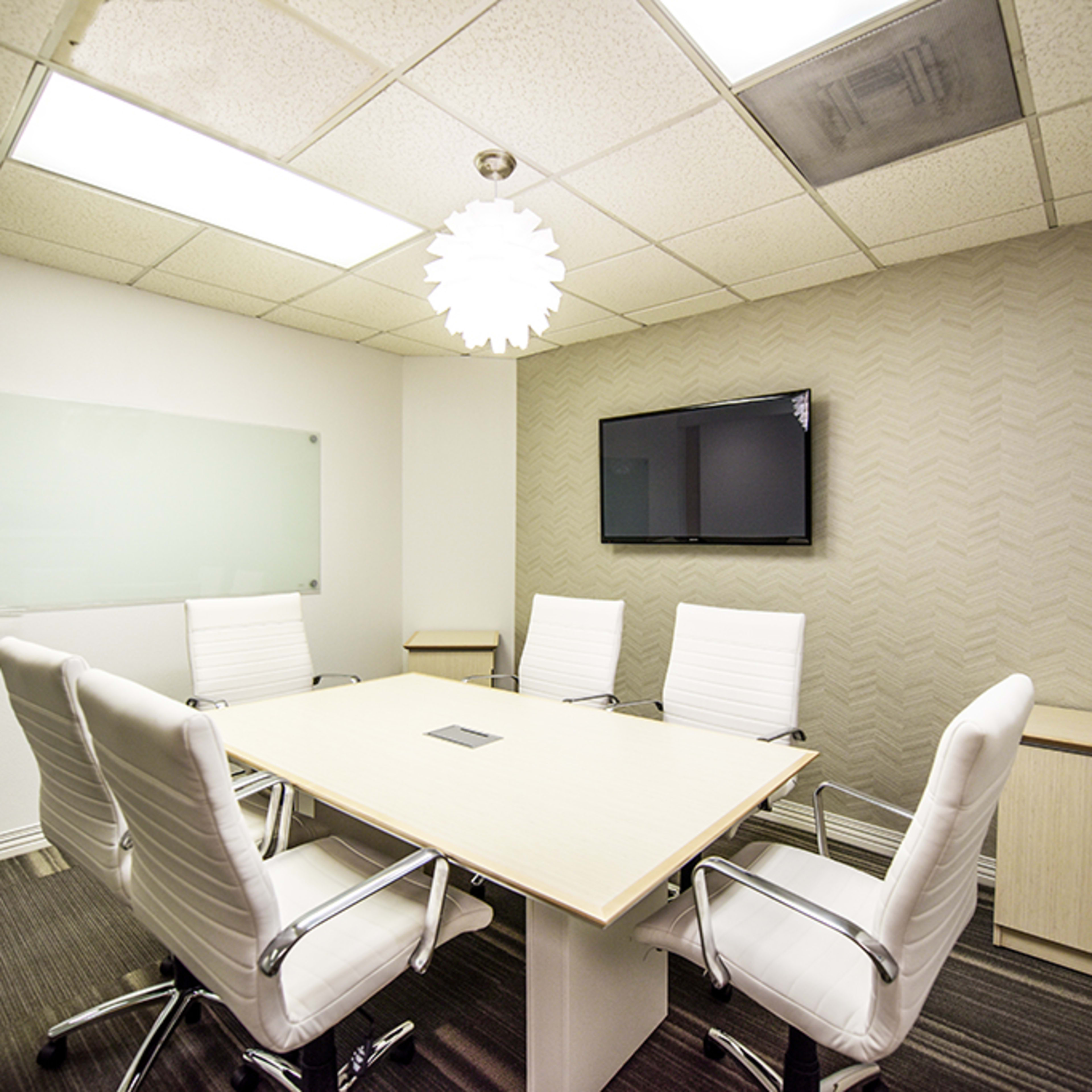 A modern conference room with a large table surrounded by white chairs, a wall-mounted television, and a decorative light fixture.
