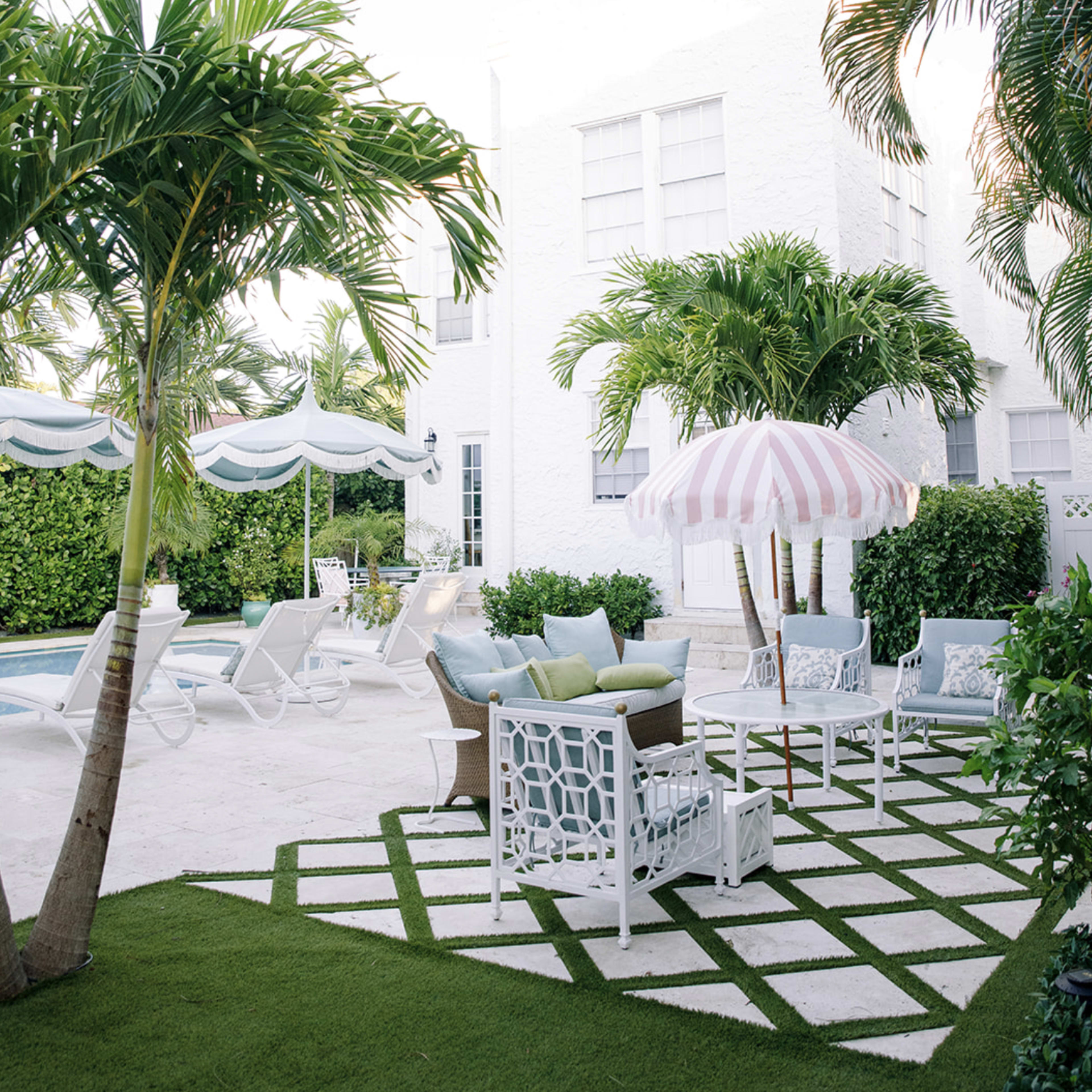 A white-walled patio features seating arrangements under a striped umbrella, surrounded by tropical plants and a pool area.