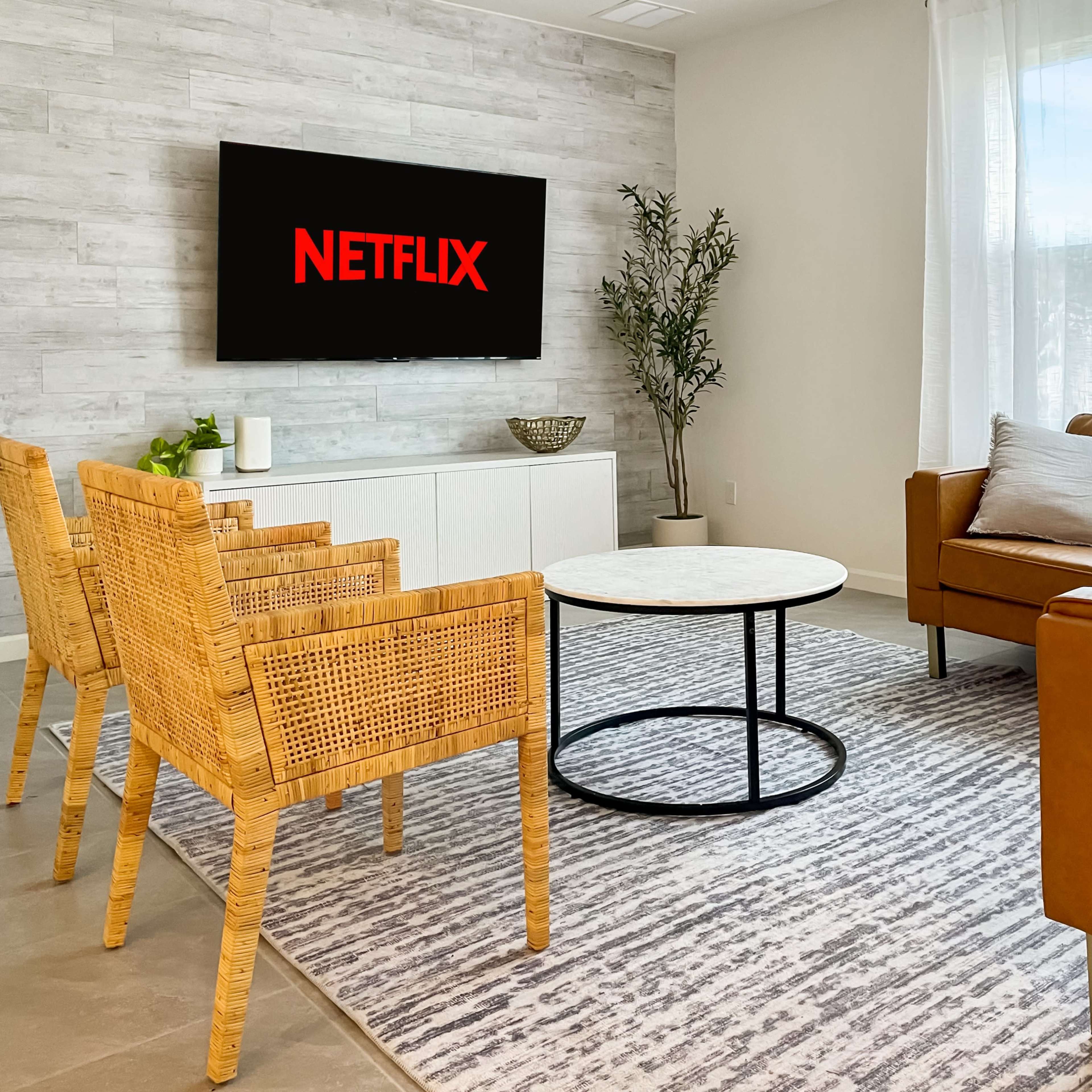 A modern living room features a television displaying the Netflix logo, a circular coffee table, and wicker chairs arranged around a light-colored rug.
