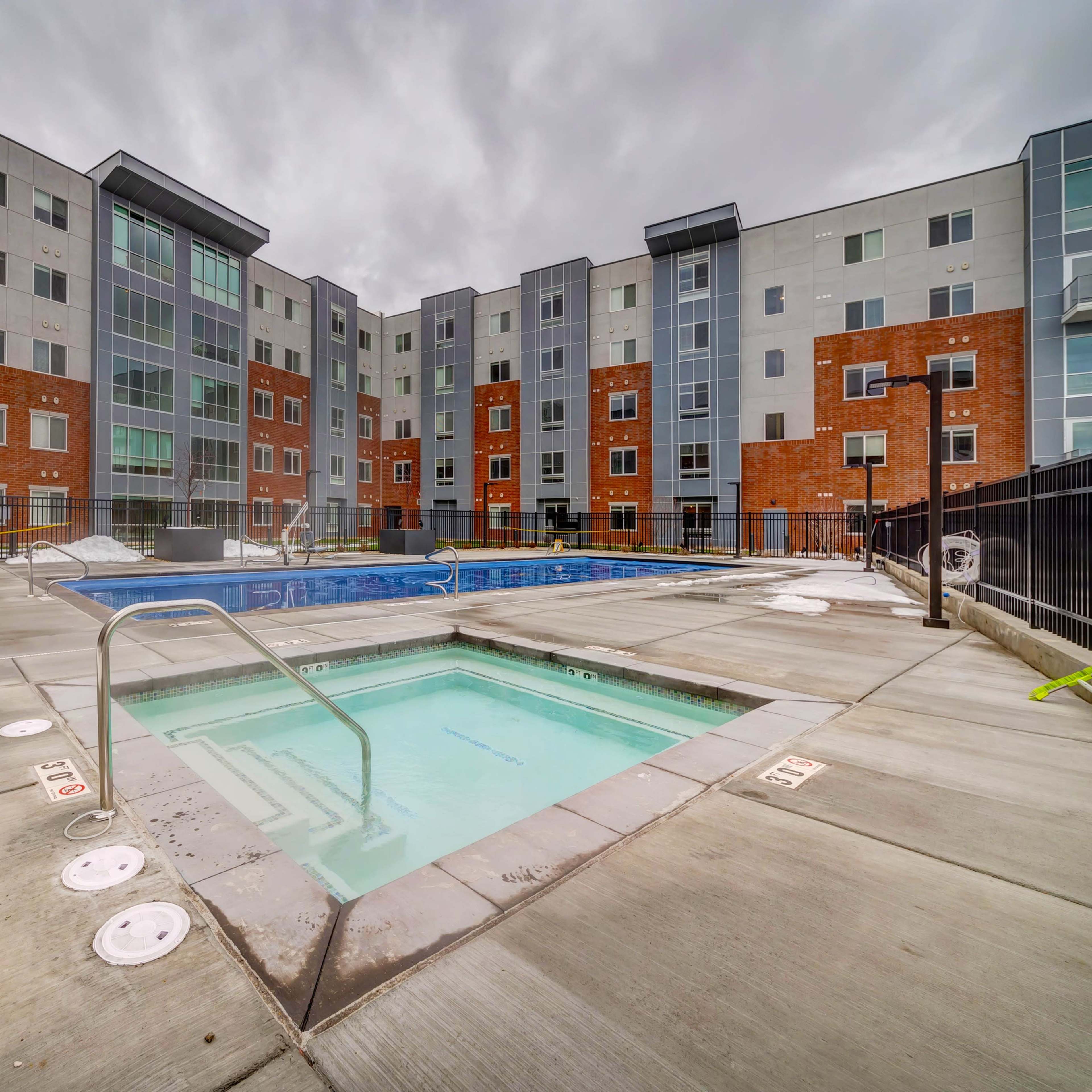 A modern apartment complex with two outdoor swimming pools, surrounded by concrete pathways and a fenced area.