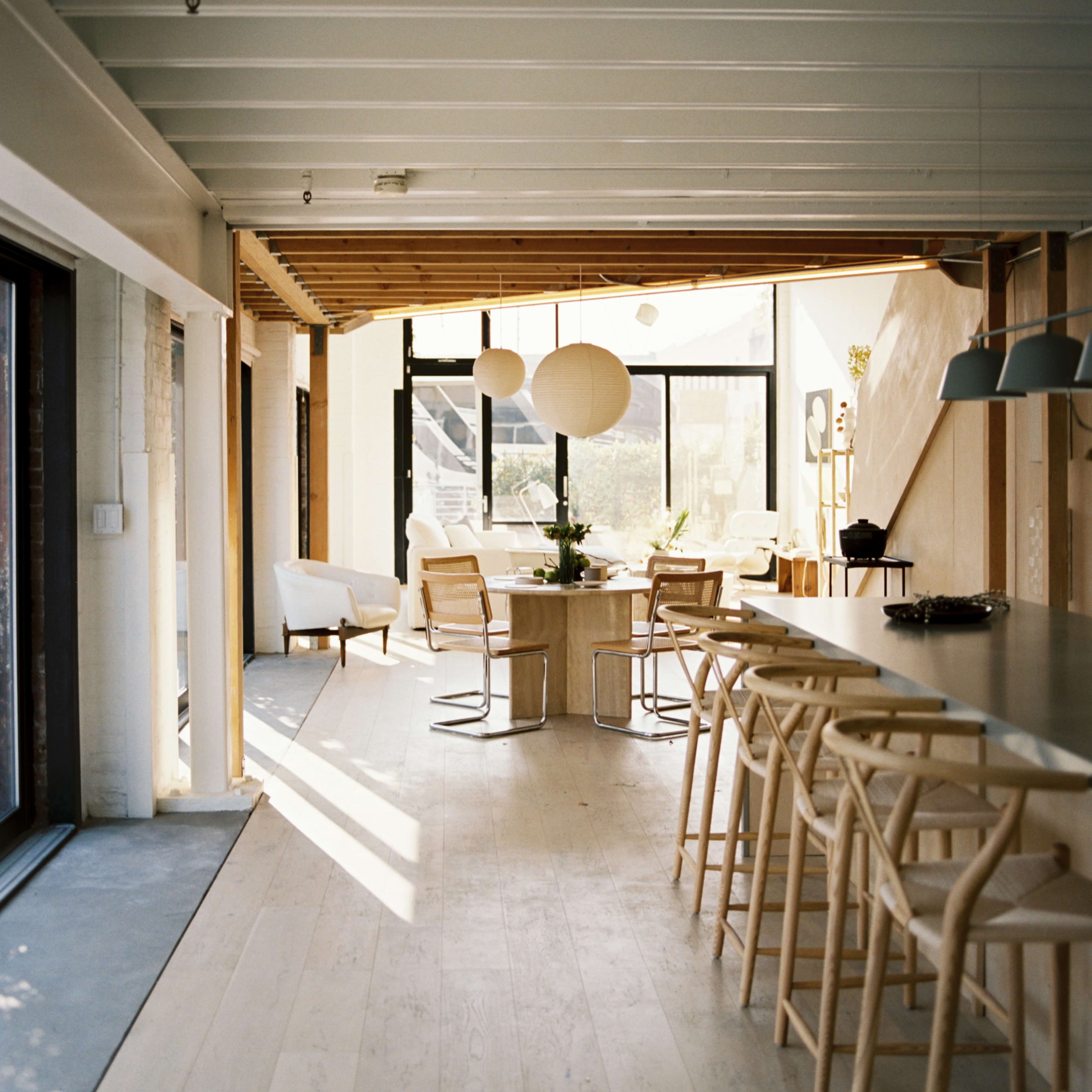 A bright, modern interior of a dining area with wooden accents, featuring a long bar with stools and a dining table surrounded by chairs, illuminated by natural light from large windows.