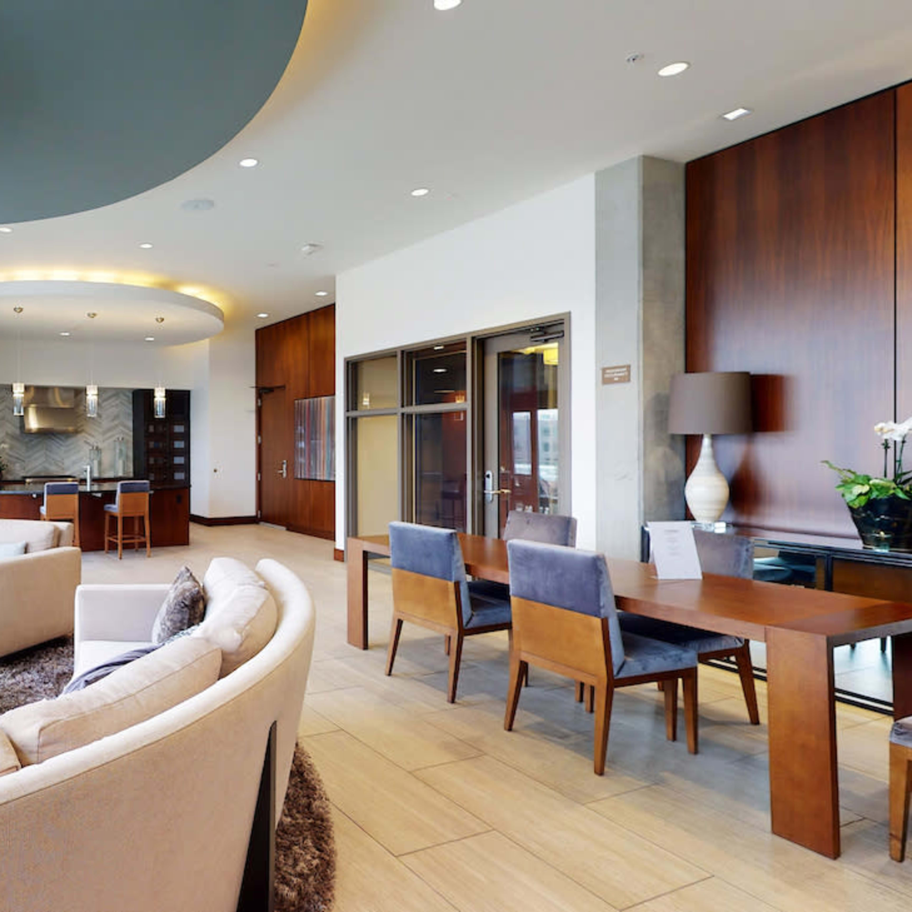 A modern, spacious living area features a large circular sofa, a dining table for four, and large windows with a view of the city.