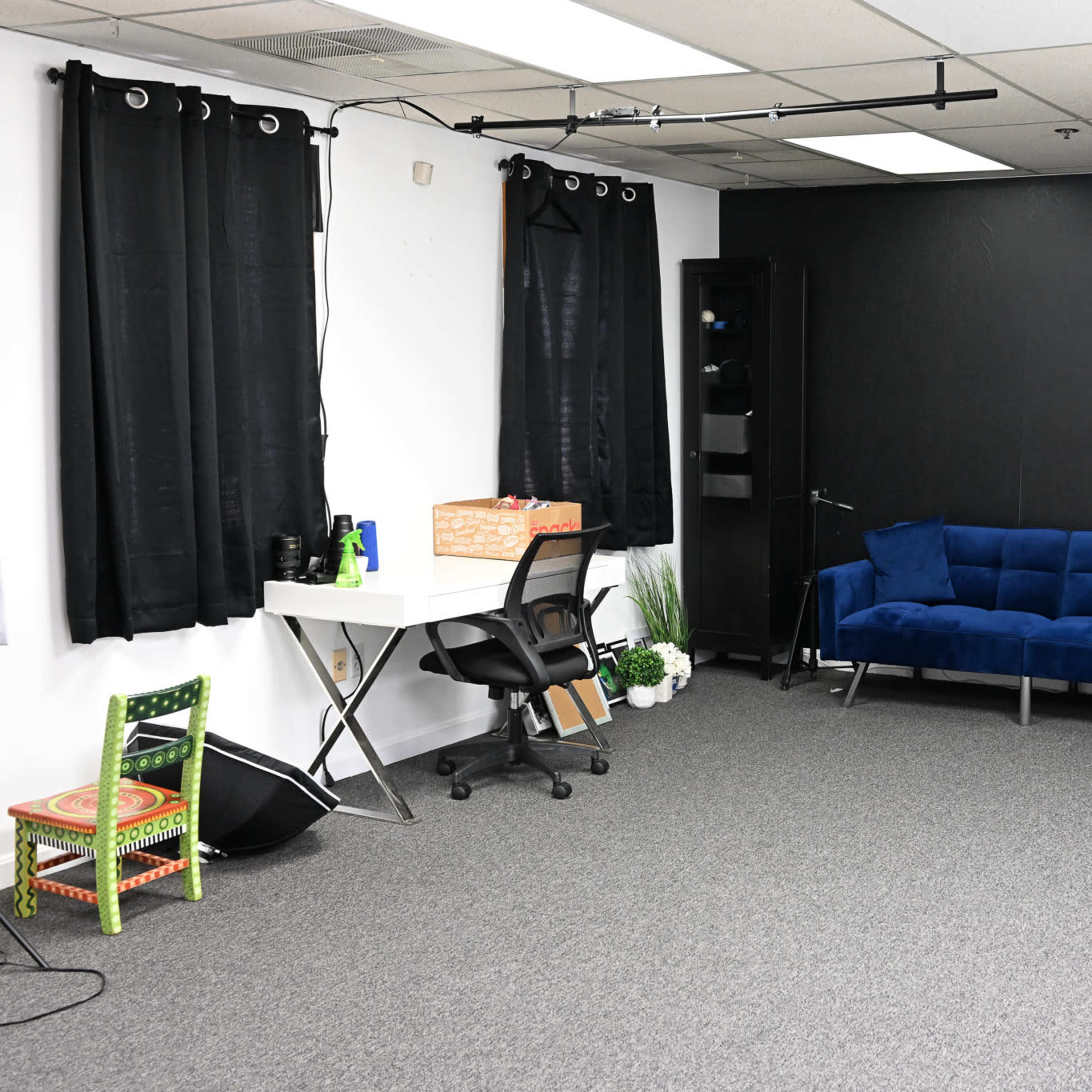 A sparse room with gray carpeting, a blue couch, a desk with a chair, two black curtains, and a few decorative plants.
