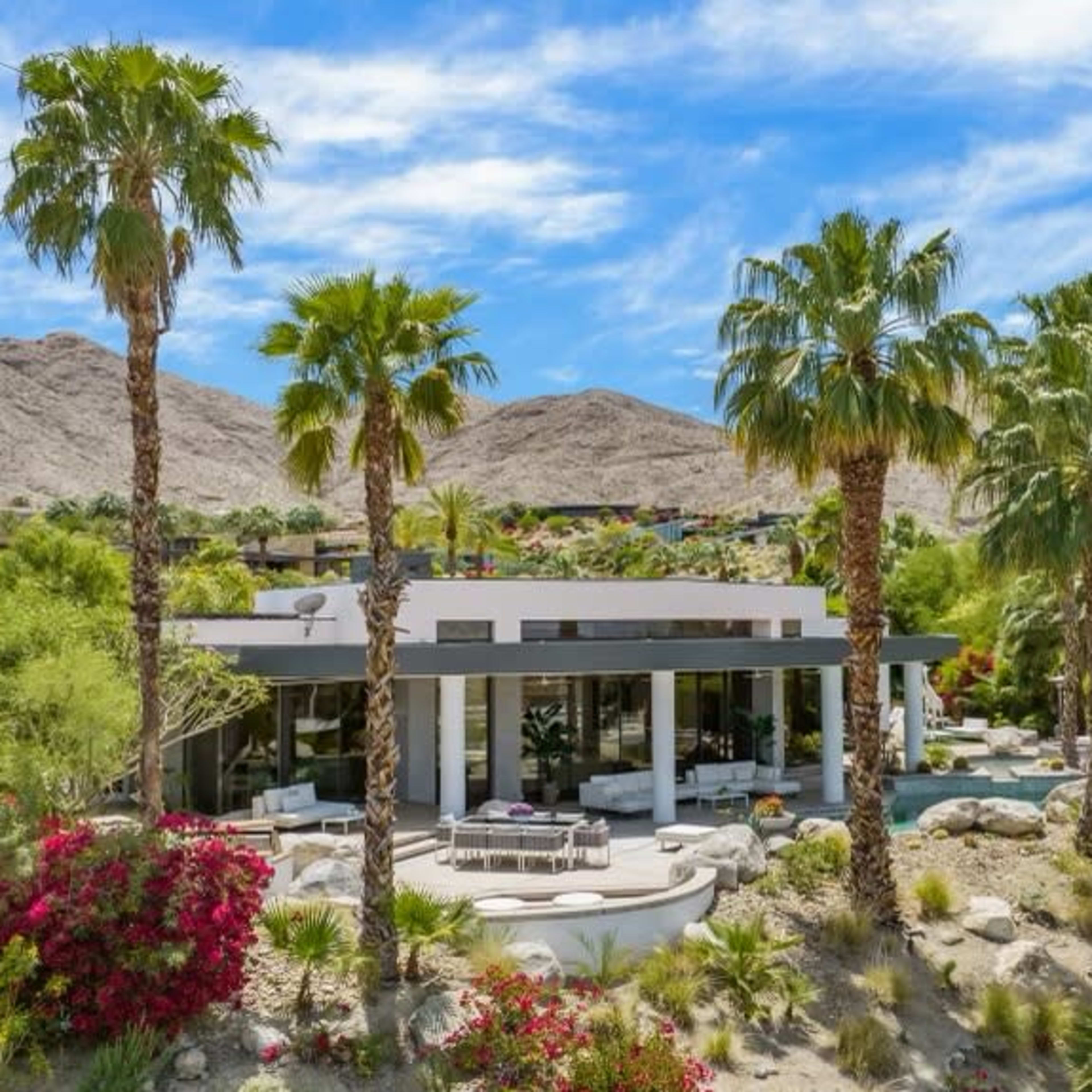 A modern house surrounded by palm trees and lush vegetation, set against a backdrop of mountains.