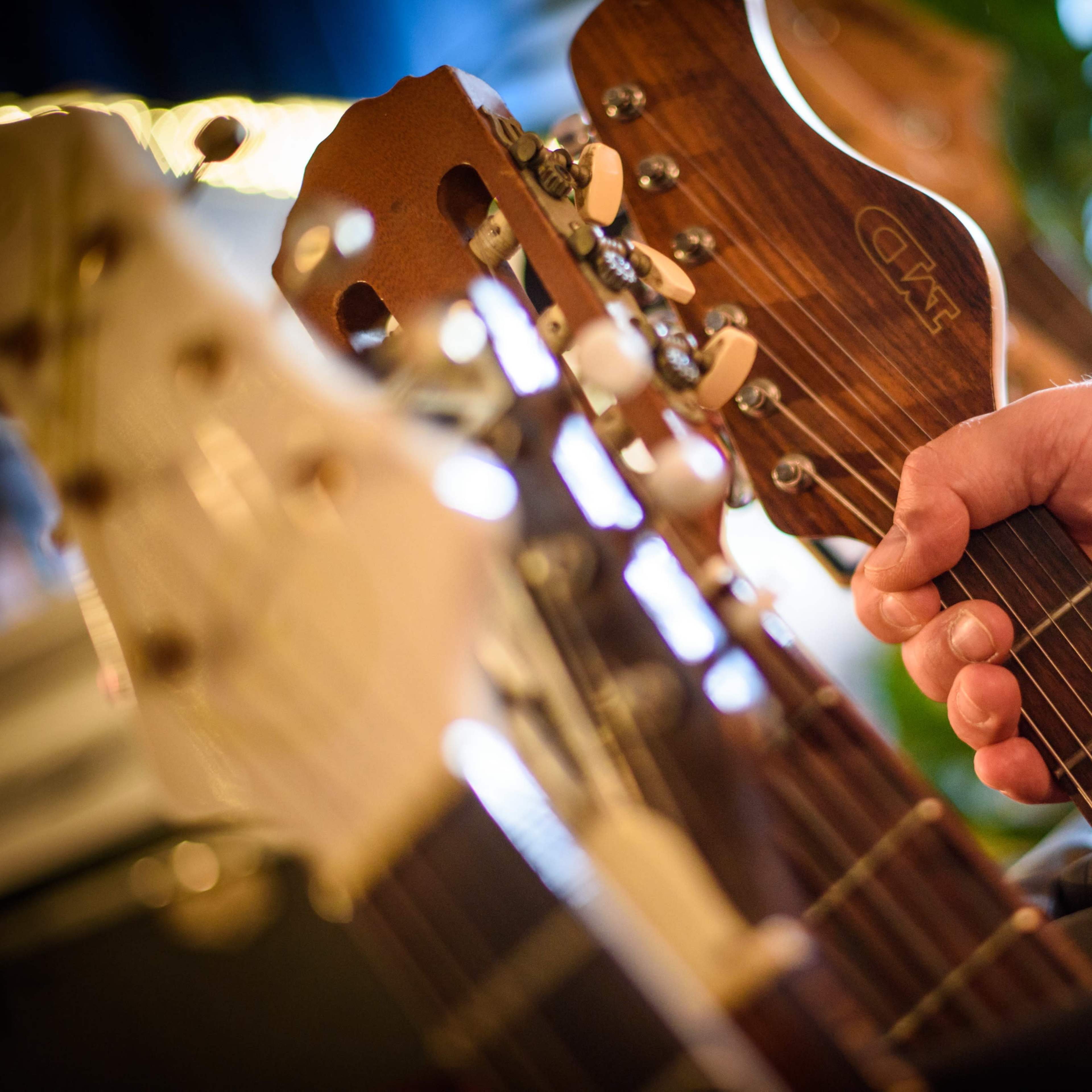 A hand grips the neck of a guitar while multiple guitars hang in the background.