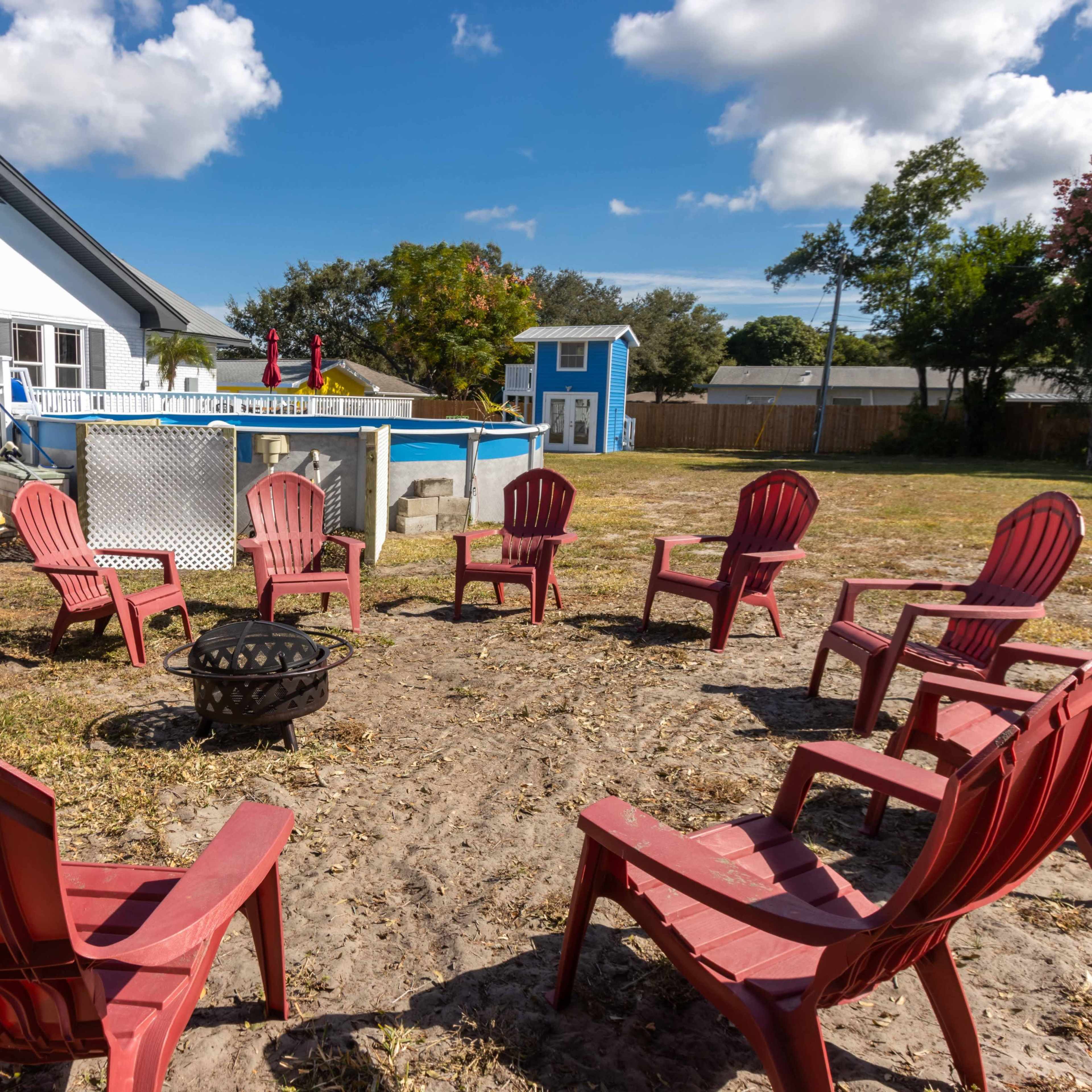 The scene shows a circular arrangement of red Adirondack chairs surrounding a fire pit in a yard beside a swimming pool and a house.