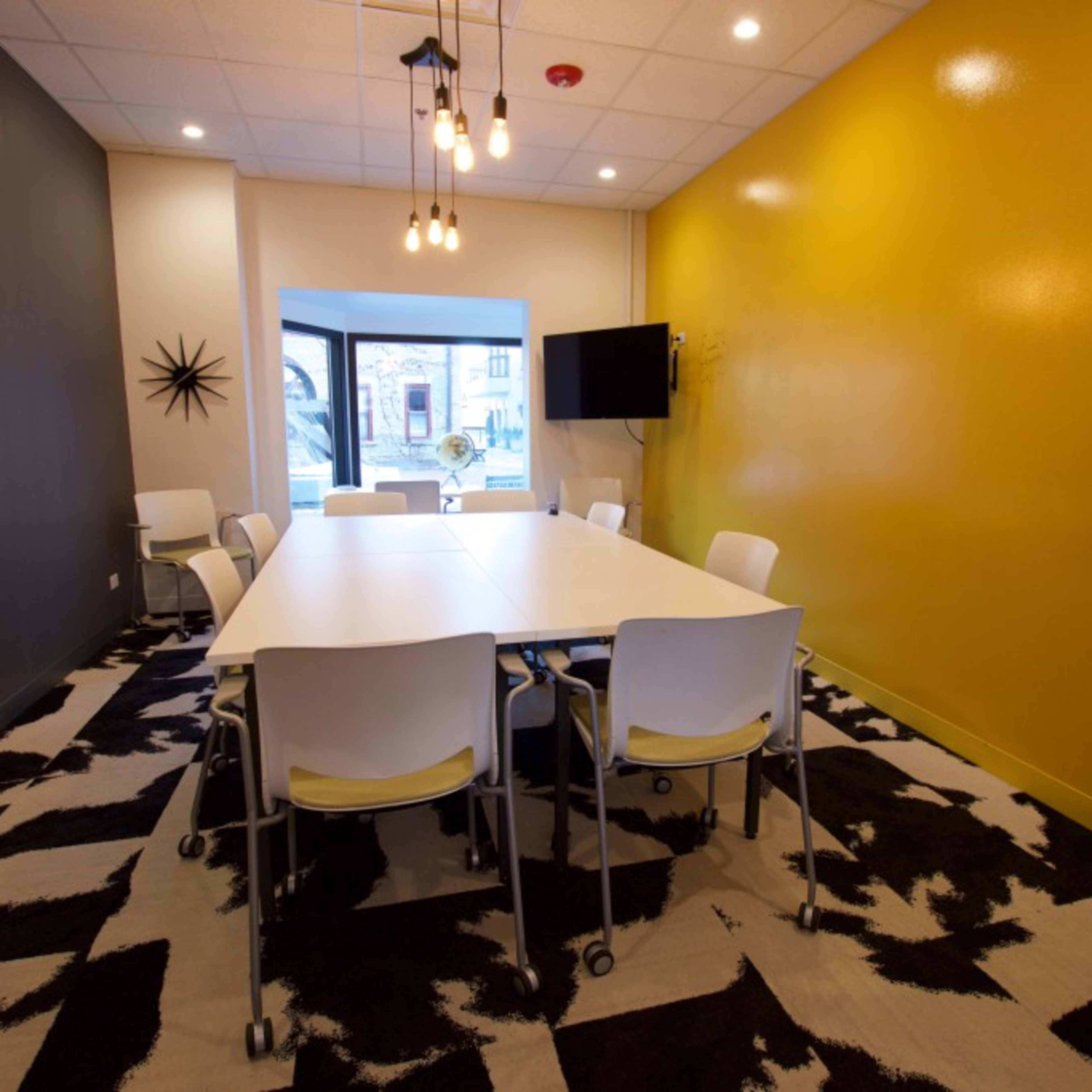 A modern meeting room features a large white table surrounded by eight rolling chairs, with a yellow accent wall and a window providing natural light.
