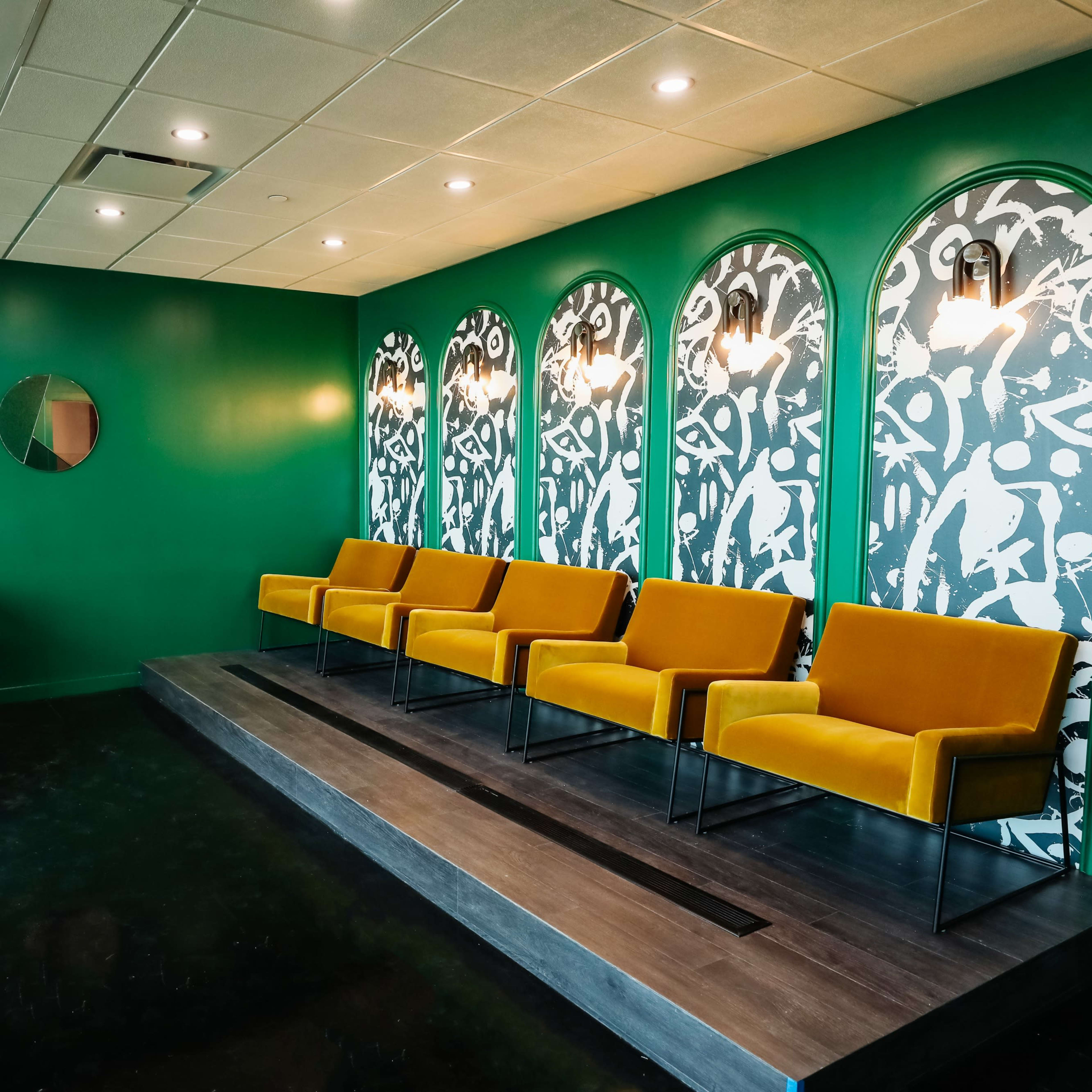 A waiting area with bright green walls, a patterned mural, four yellow chairs, and a bar with stools against one wall.