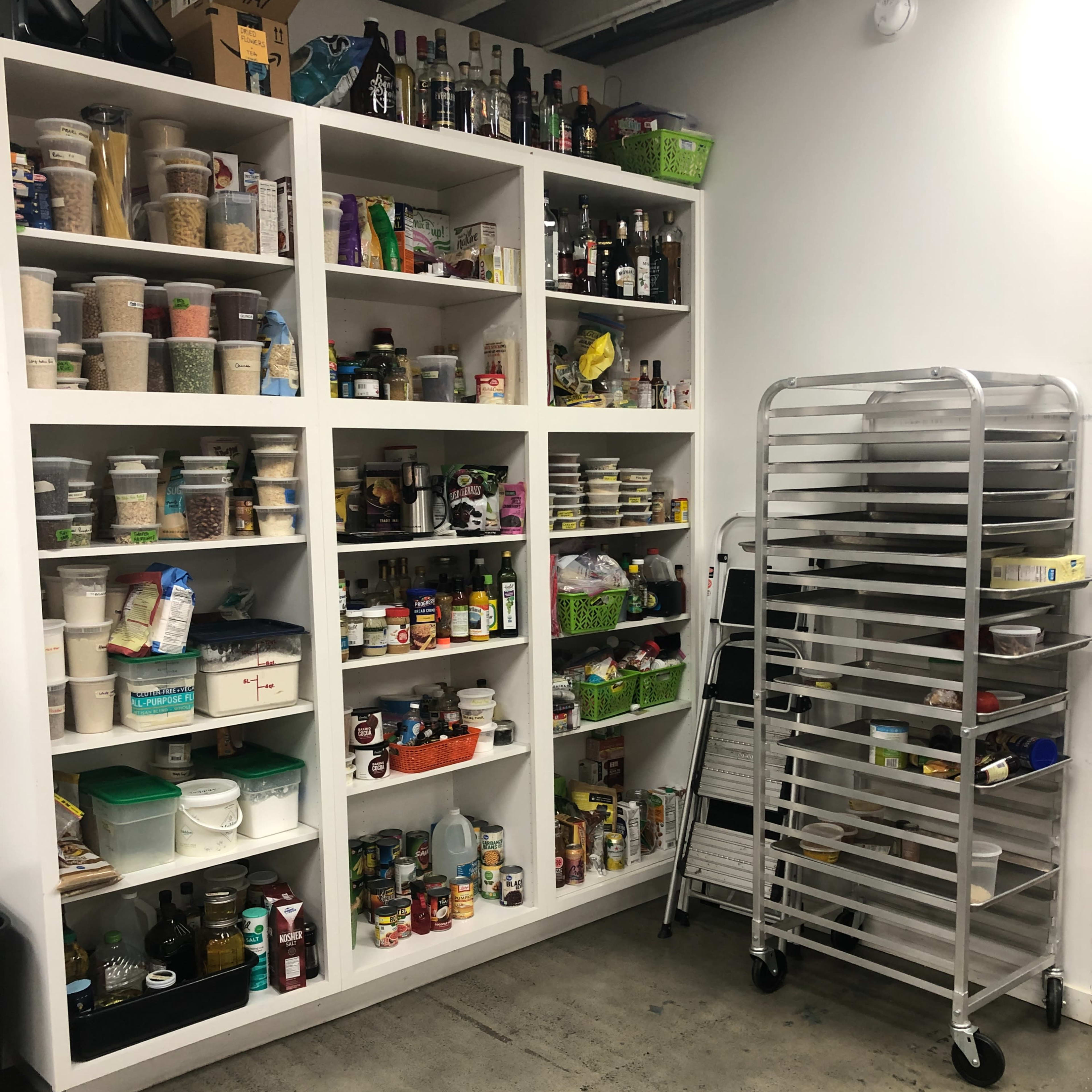 A storage area with shelves filled with various jars, cans, and bottles, alongside a mobile rack in a corner.