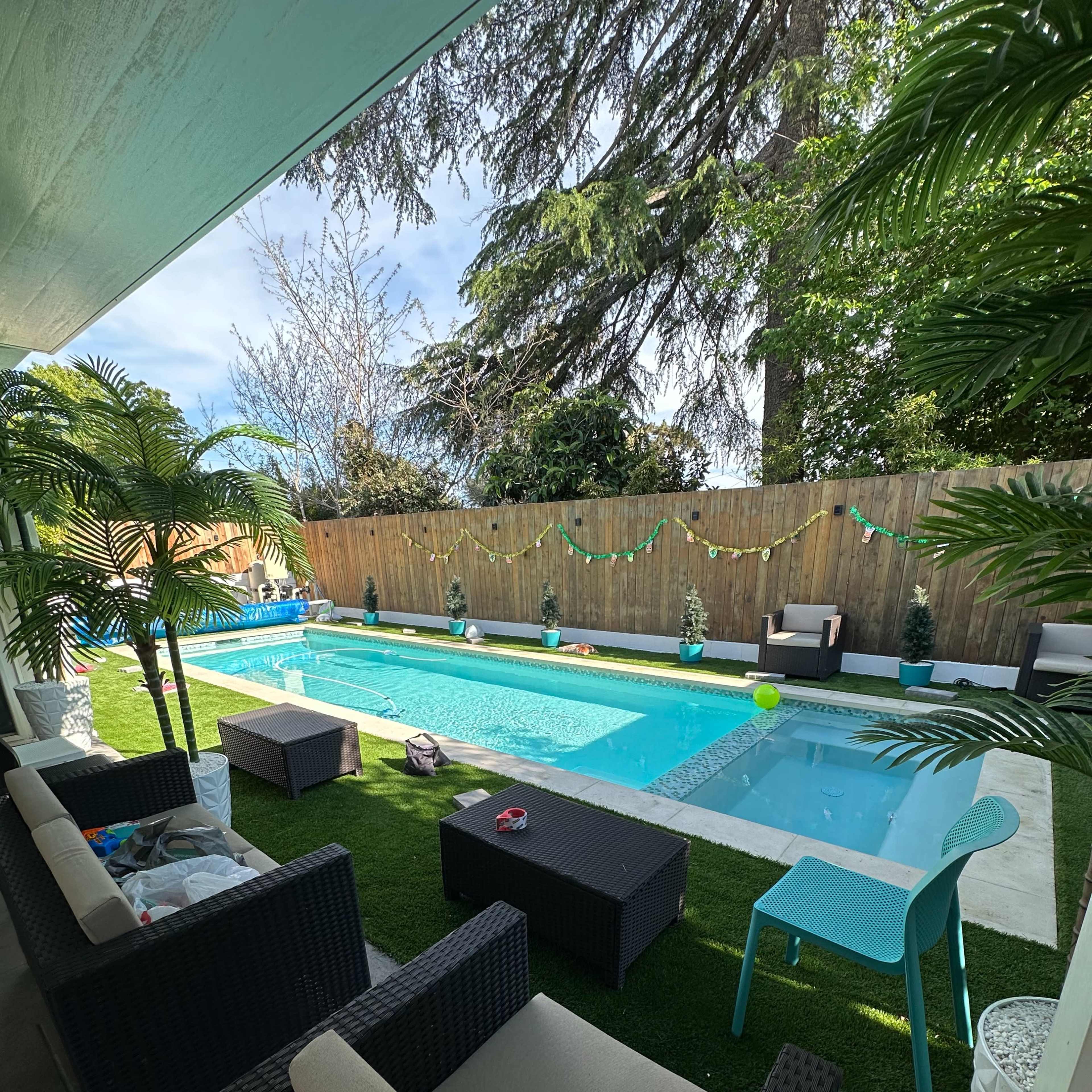 A backyard with a swimming pool surrounded by potted plants and seating areas on a grassy lawn.