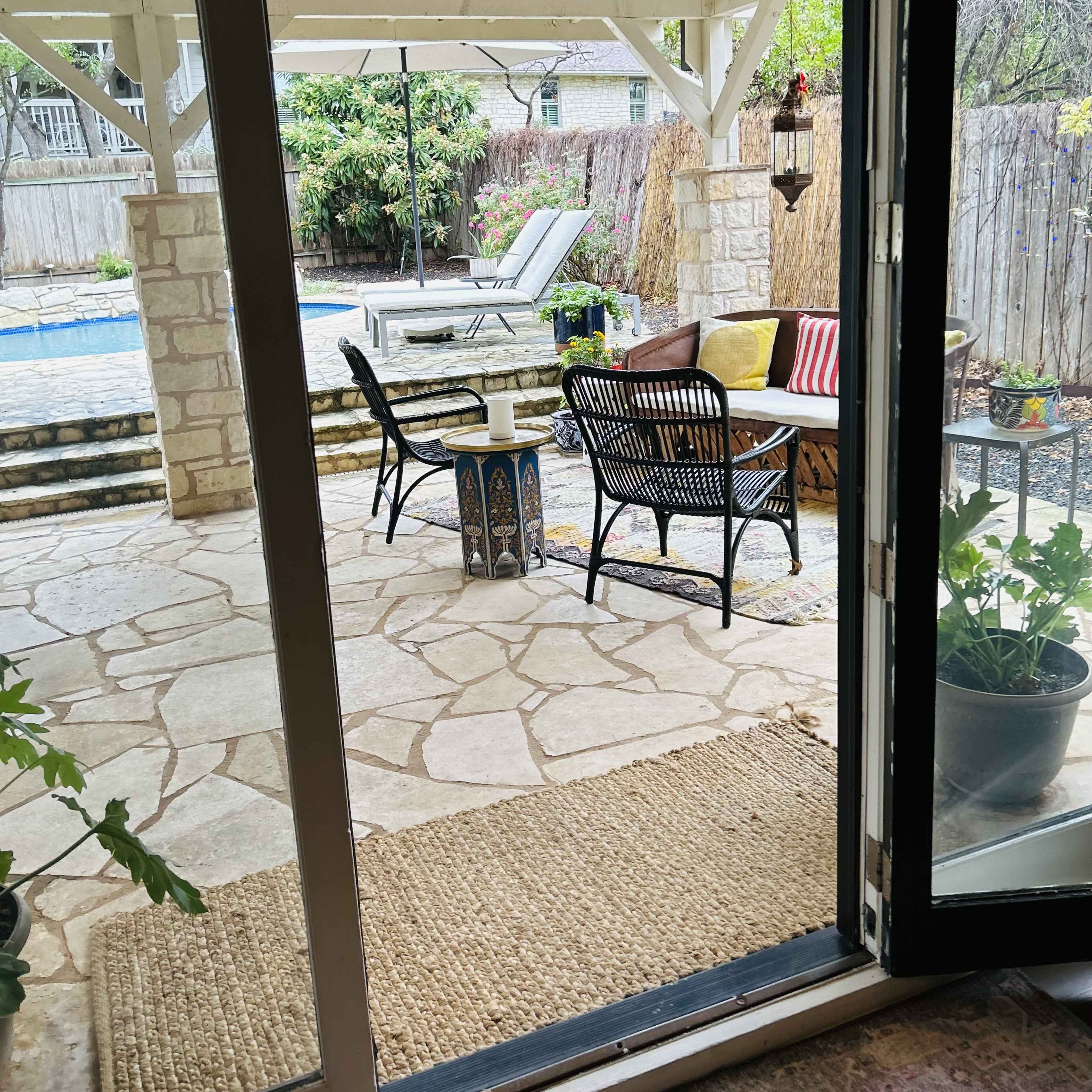 A cozy outdoor seating area with stone flooring, featuring a small table and chairs, overlooking a pool surrounded by greenery.