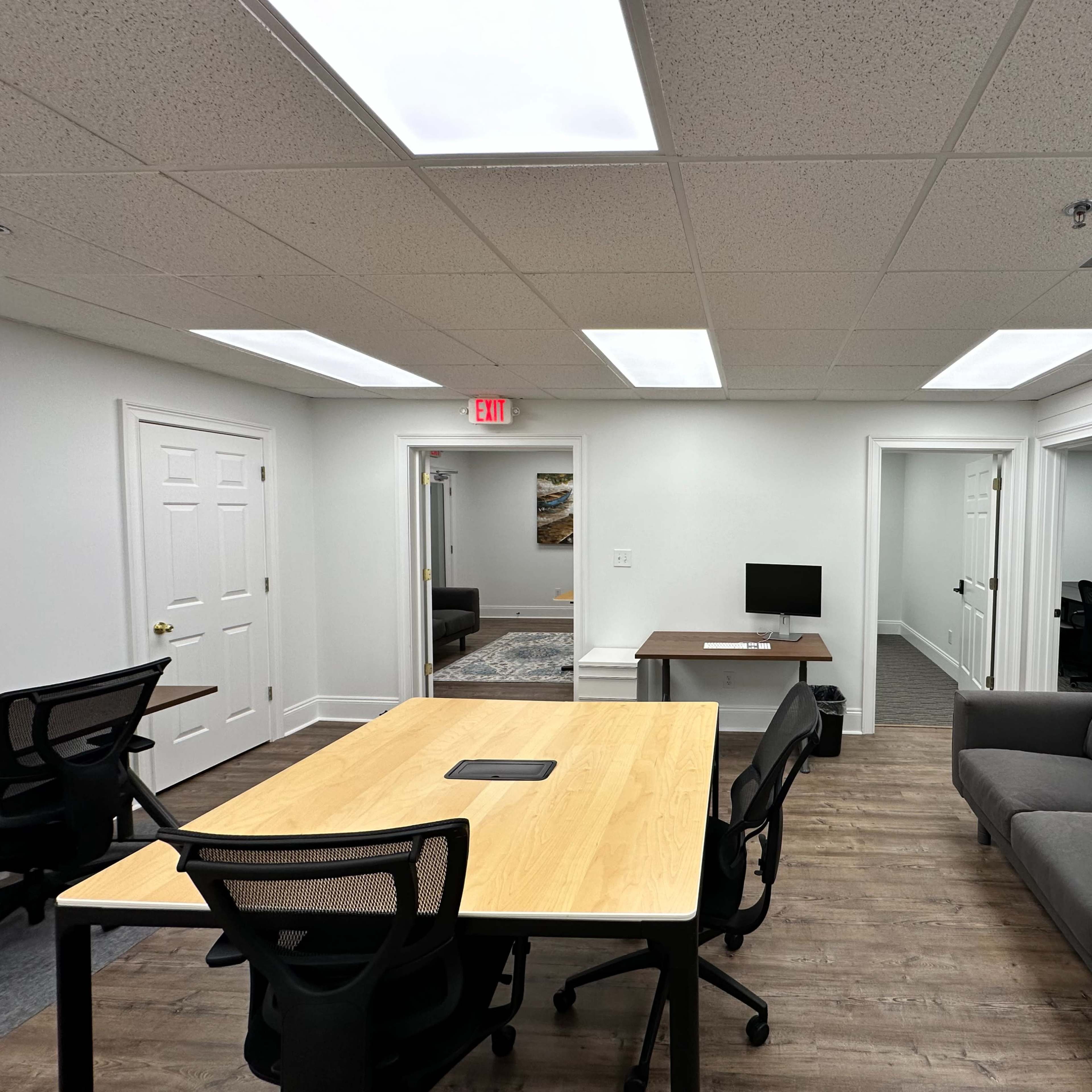 A modern office space with a wooden table, black chairs, a computer on a desk, and a gray sofa.