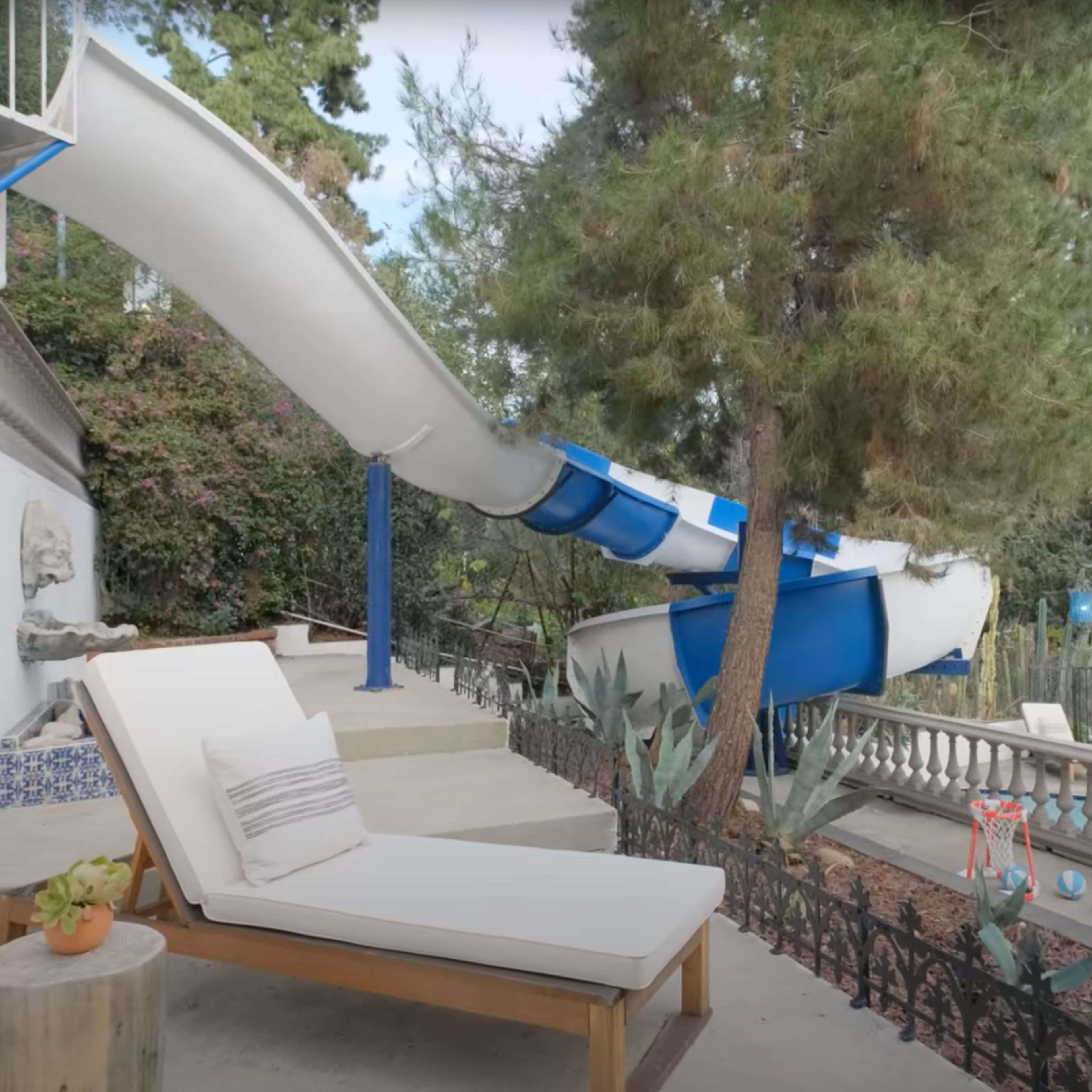 A curved blue water slide extends from a raised platform next to a sunbed, surrounded by pine trees and a fenced area with lounge chairs and a swimming pool in the background.