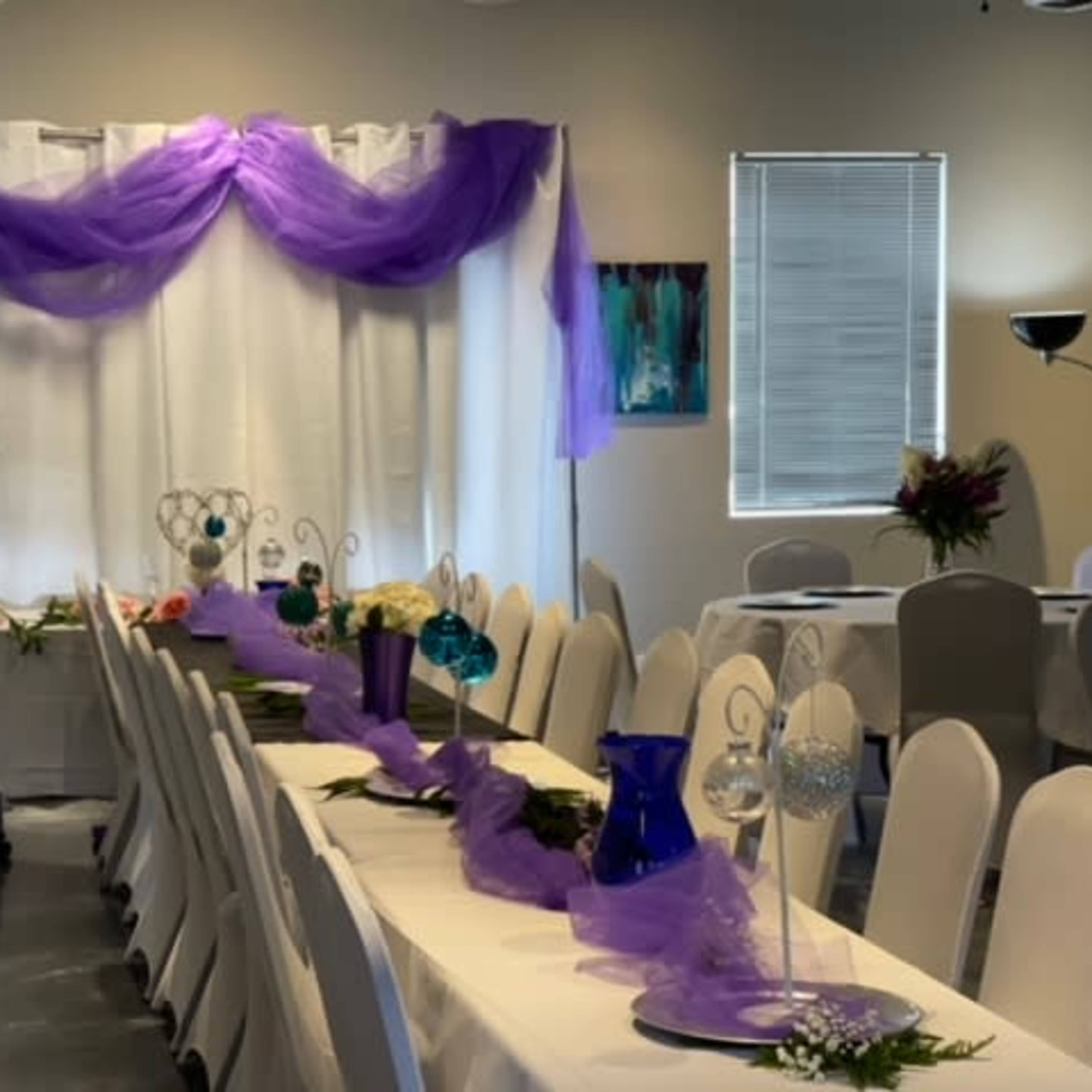 A dining area is arranged with white tables and chairs, adorned with purple drapery and decorative centerpieces.