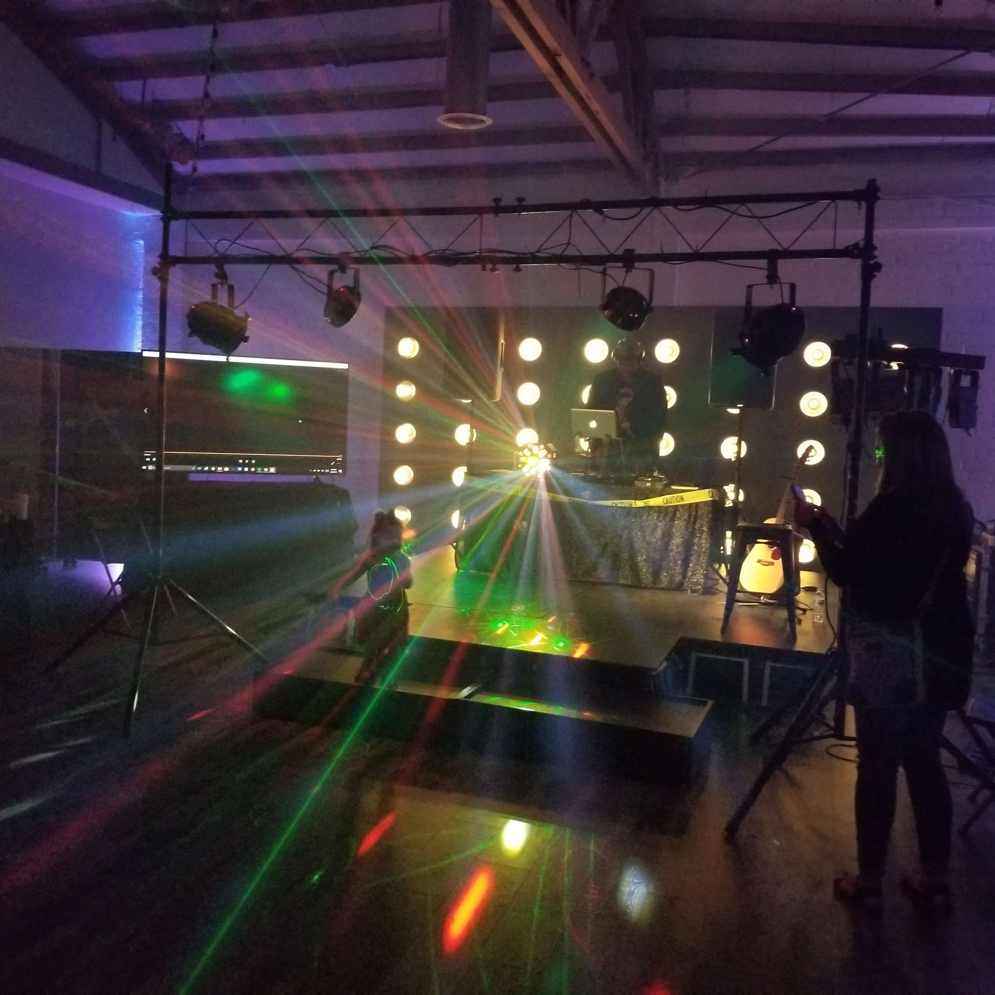 A dimly lit event space with colorful lights and a DJ setup facing a small stage.
