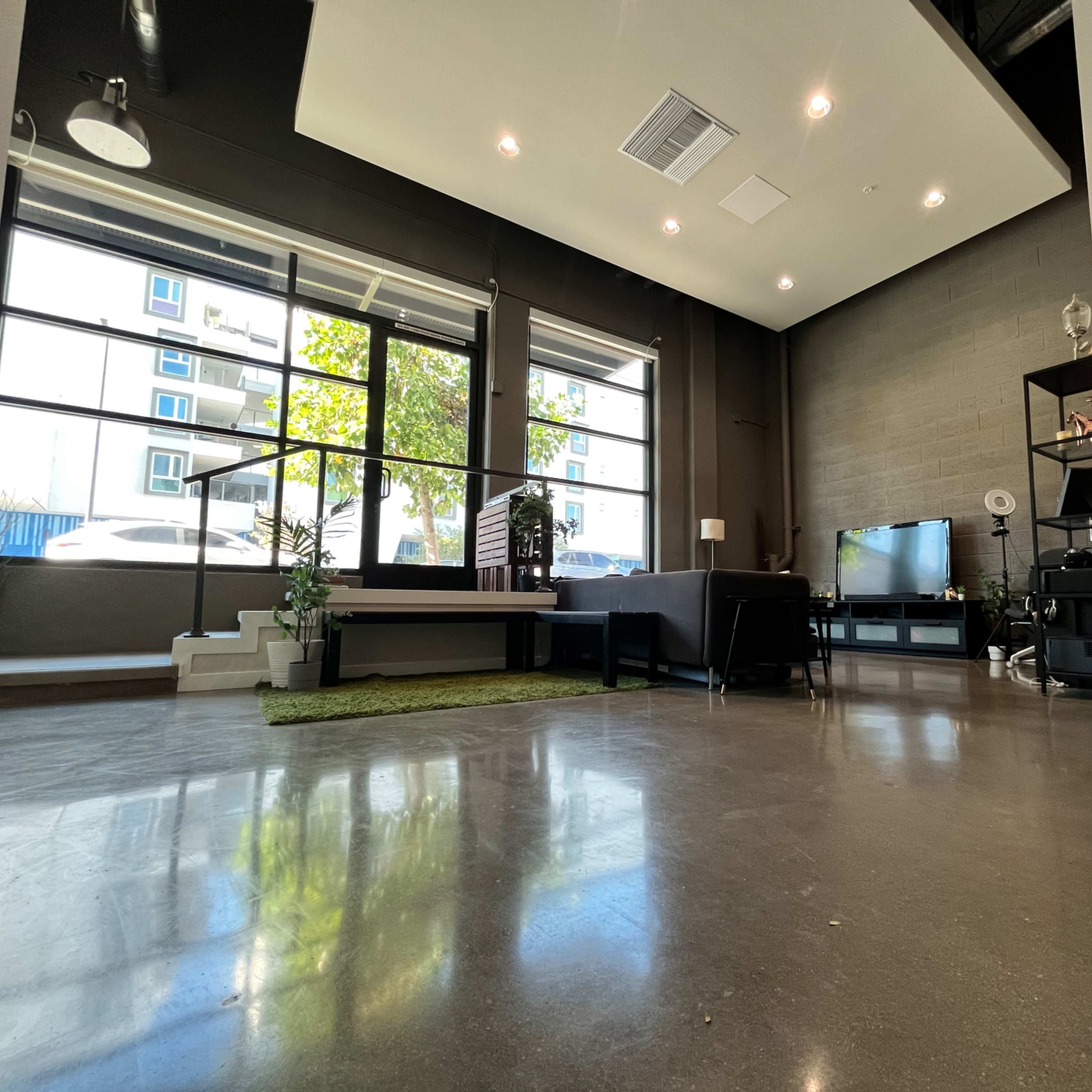 A spacious interior with large windows, a sleek concrete floor, and modern furniture arranged around a TV and a table.