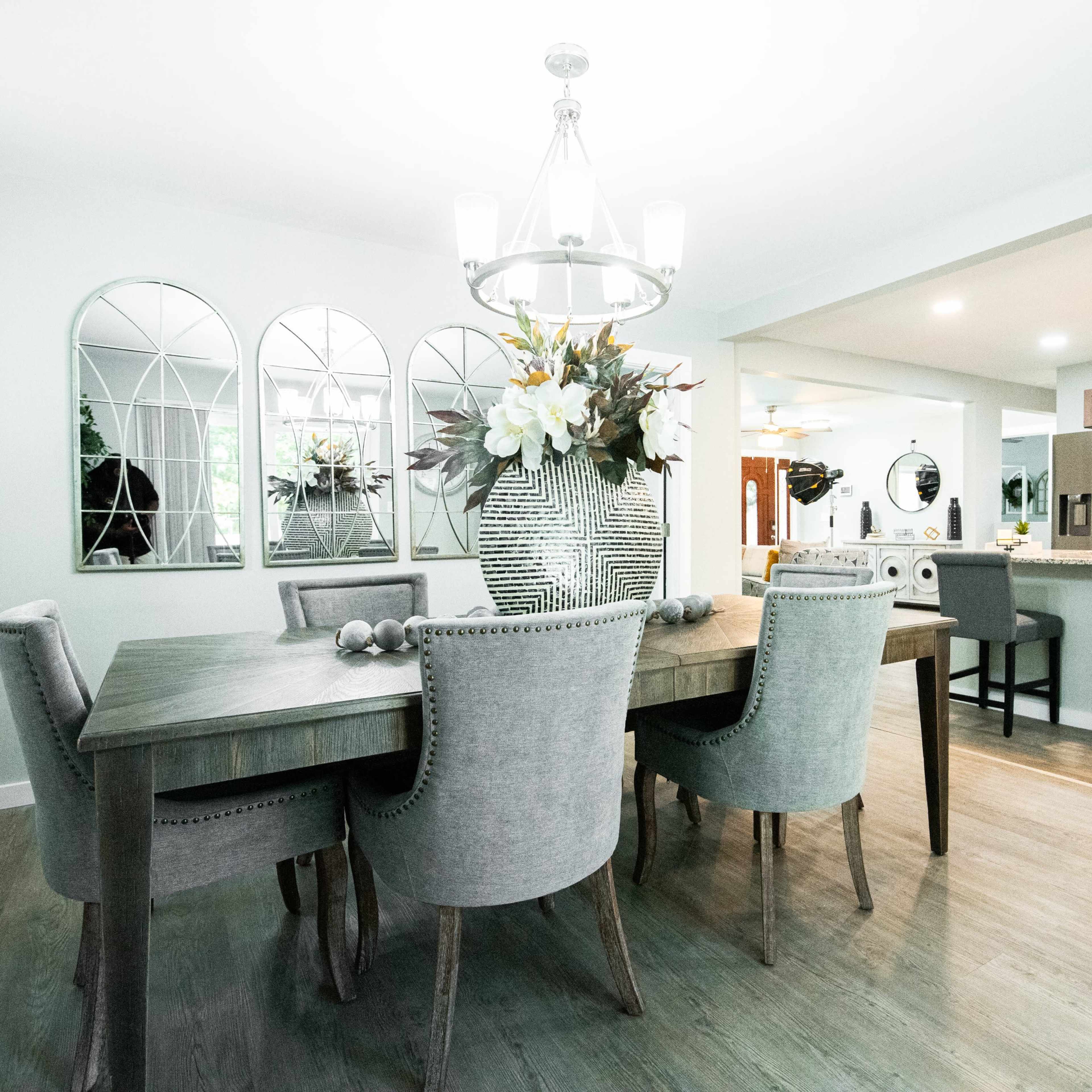 A dining room features a wooden table surrounded by upholstered chairs, with a chandelier above and decorative mirrors on the wall.