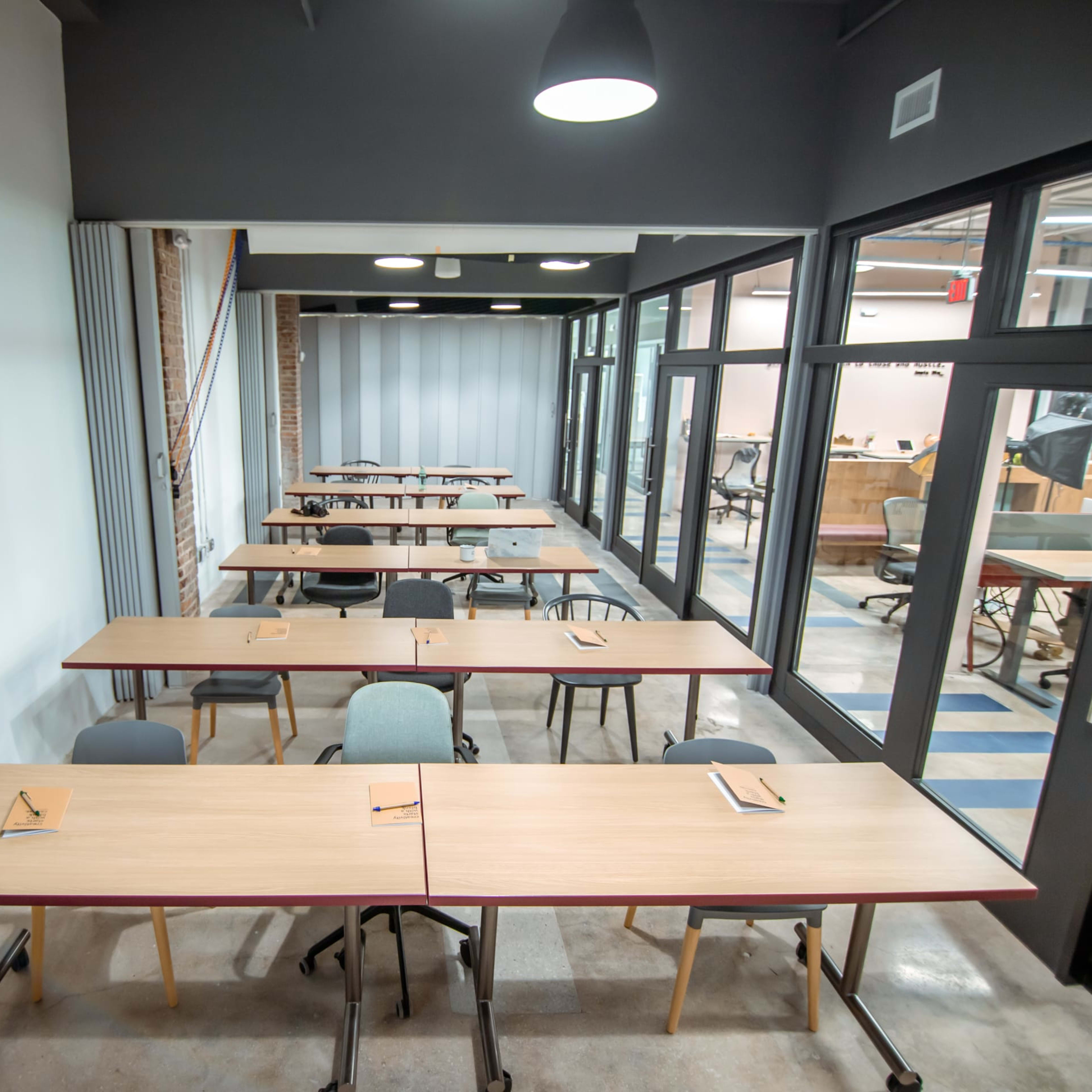 A spacious classroom with rectangular wooden tables arranged in rows, each set with a piece of paper and a pencil.