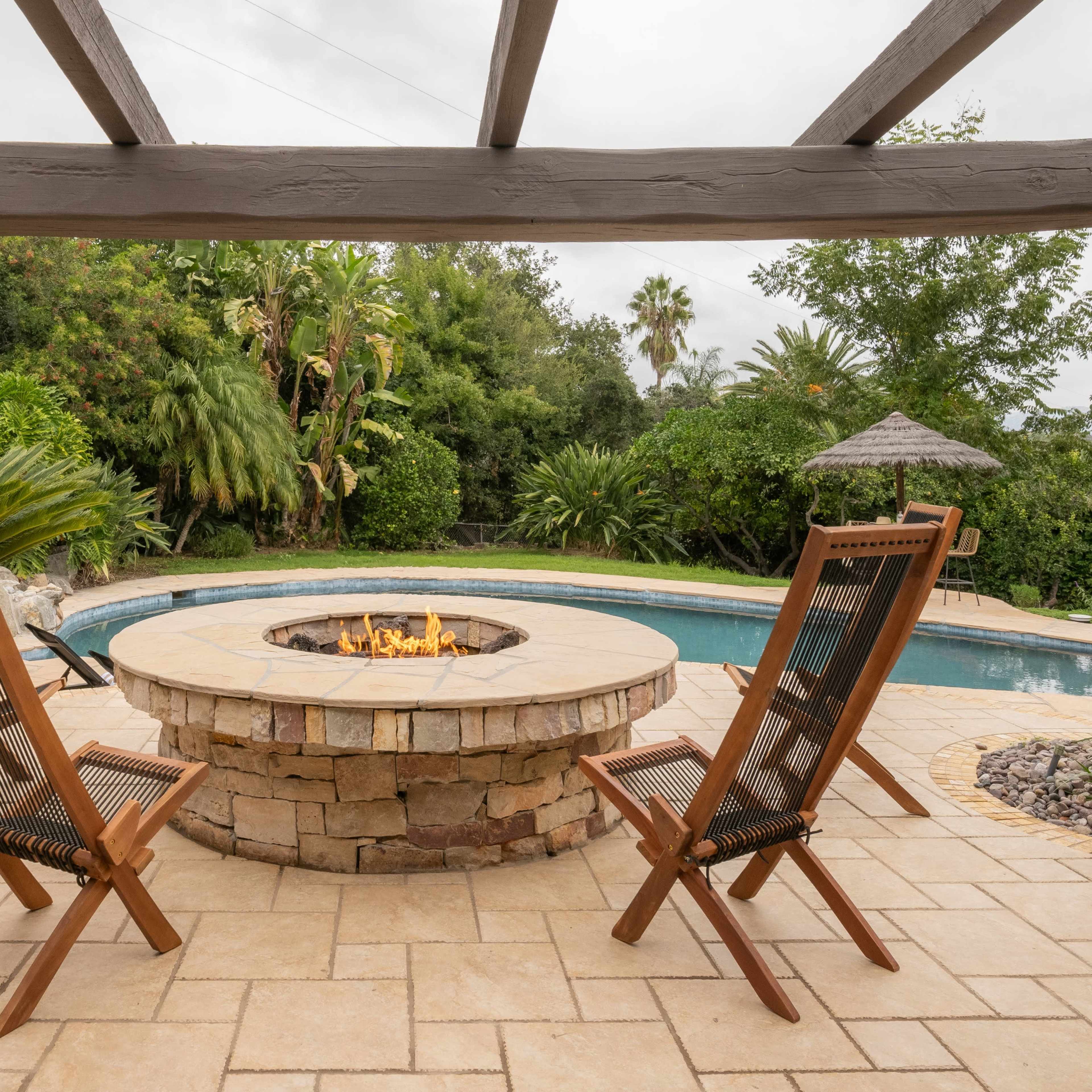A stone fire pit surrounded by two wooden chairs, overlooking a pool and lush greenery in a residential backyard.