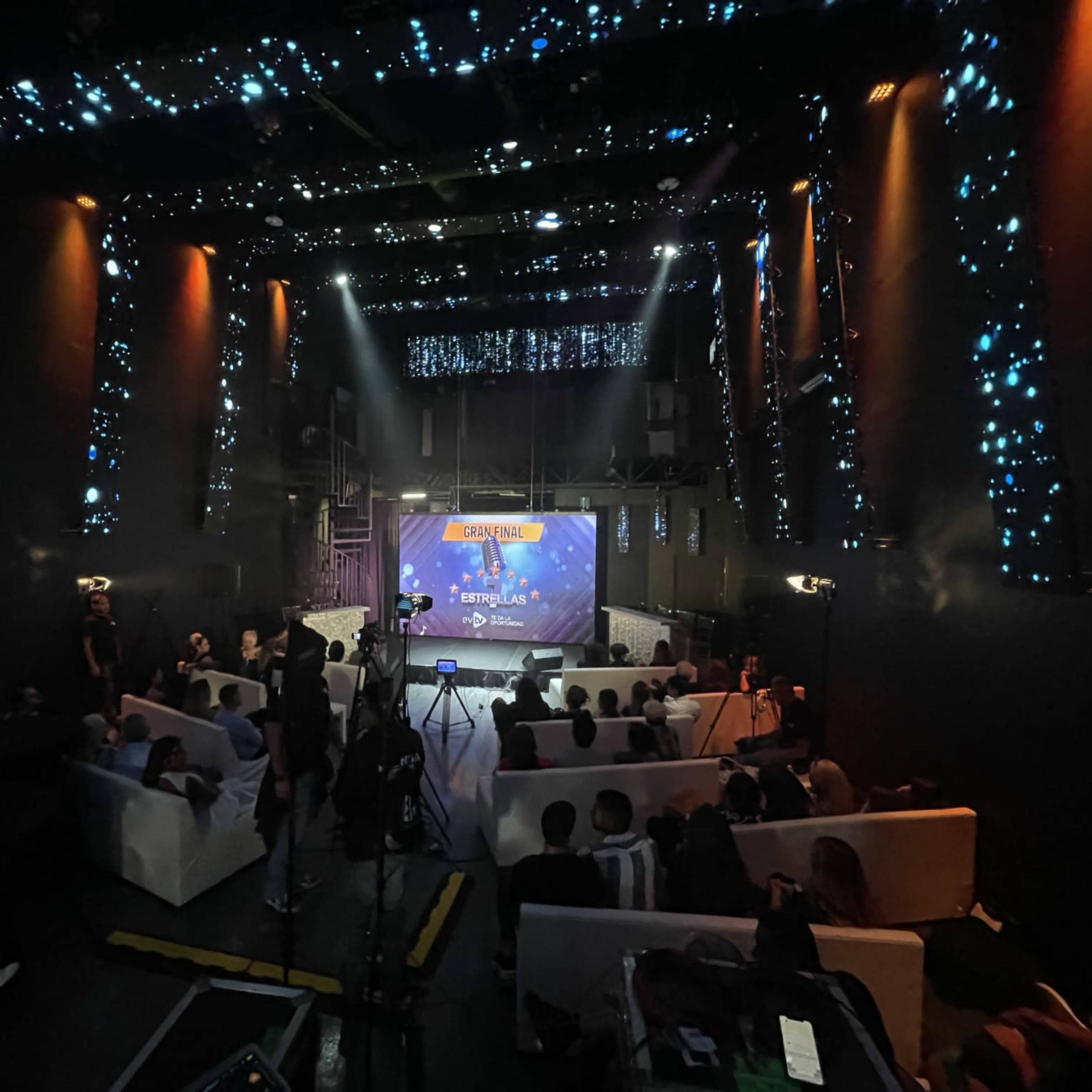 A dimly lit event space featuring a stage with bright lights and a screen, surrounded by seated attendees.