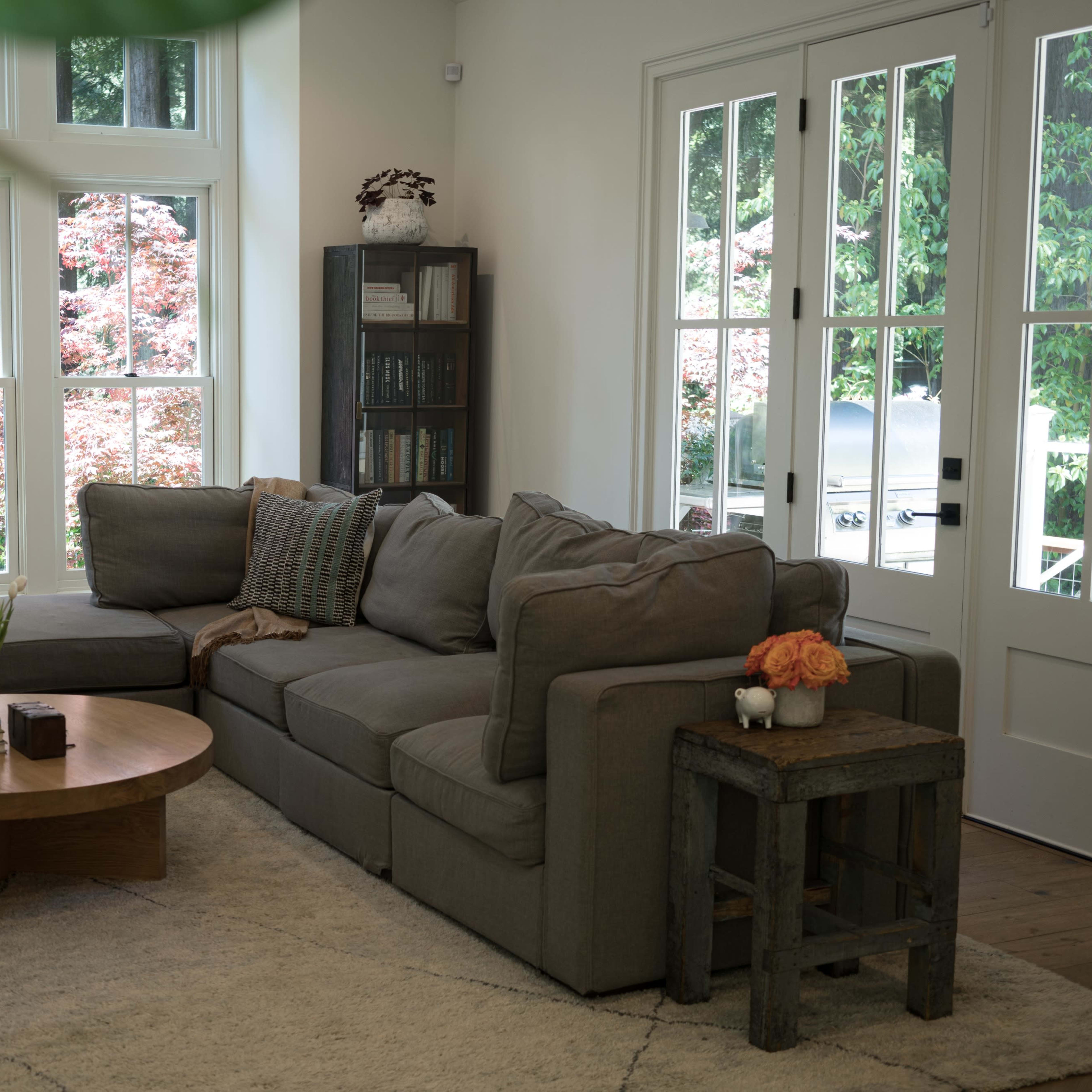 A modern living room features a large sectional sofa, a round wooden coffee table, and large windows showcasing a garden view.