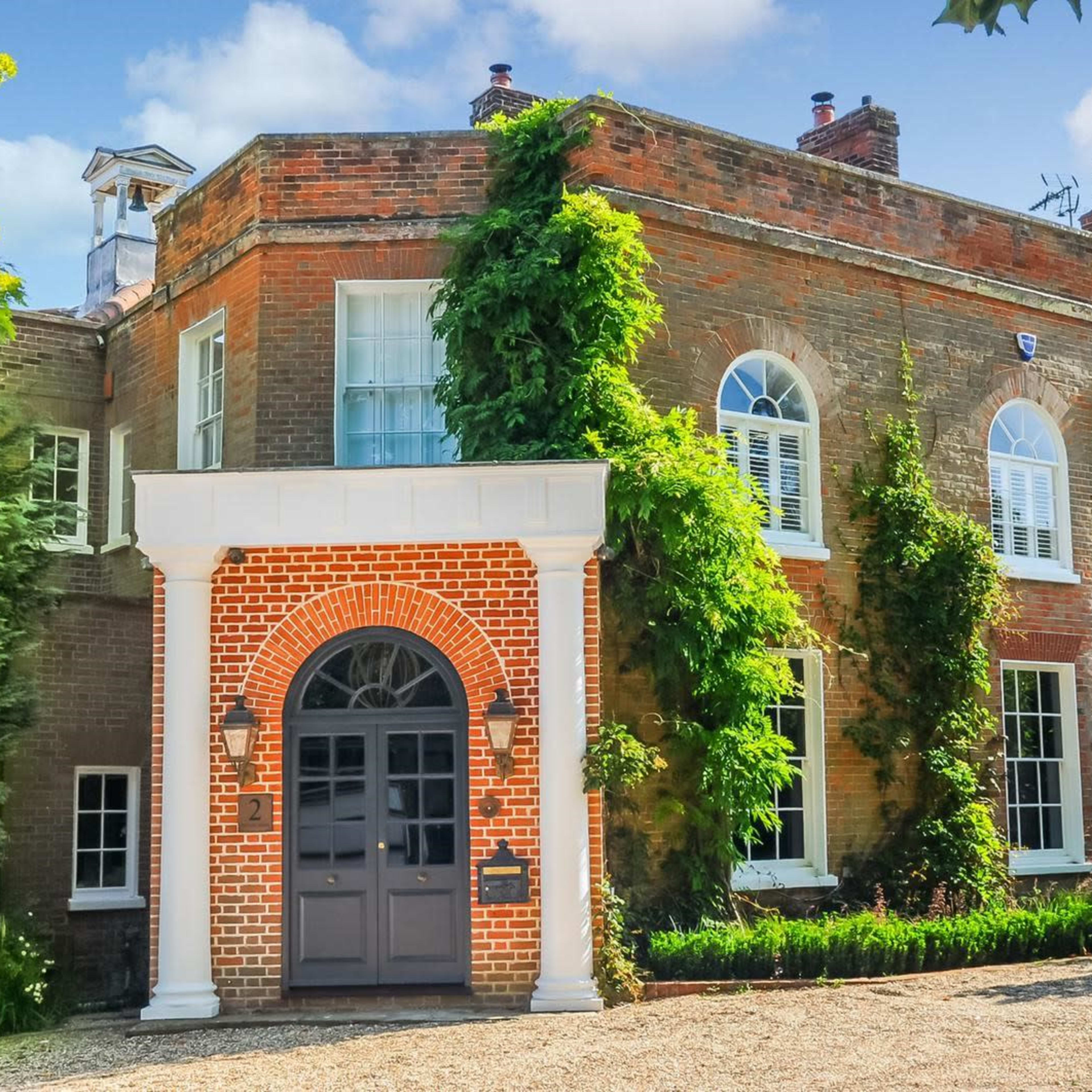 A two-story brick house with large windows and a front entrance framed by decorative columns is surrounded by greenery.