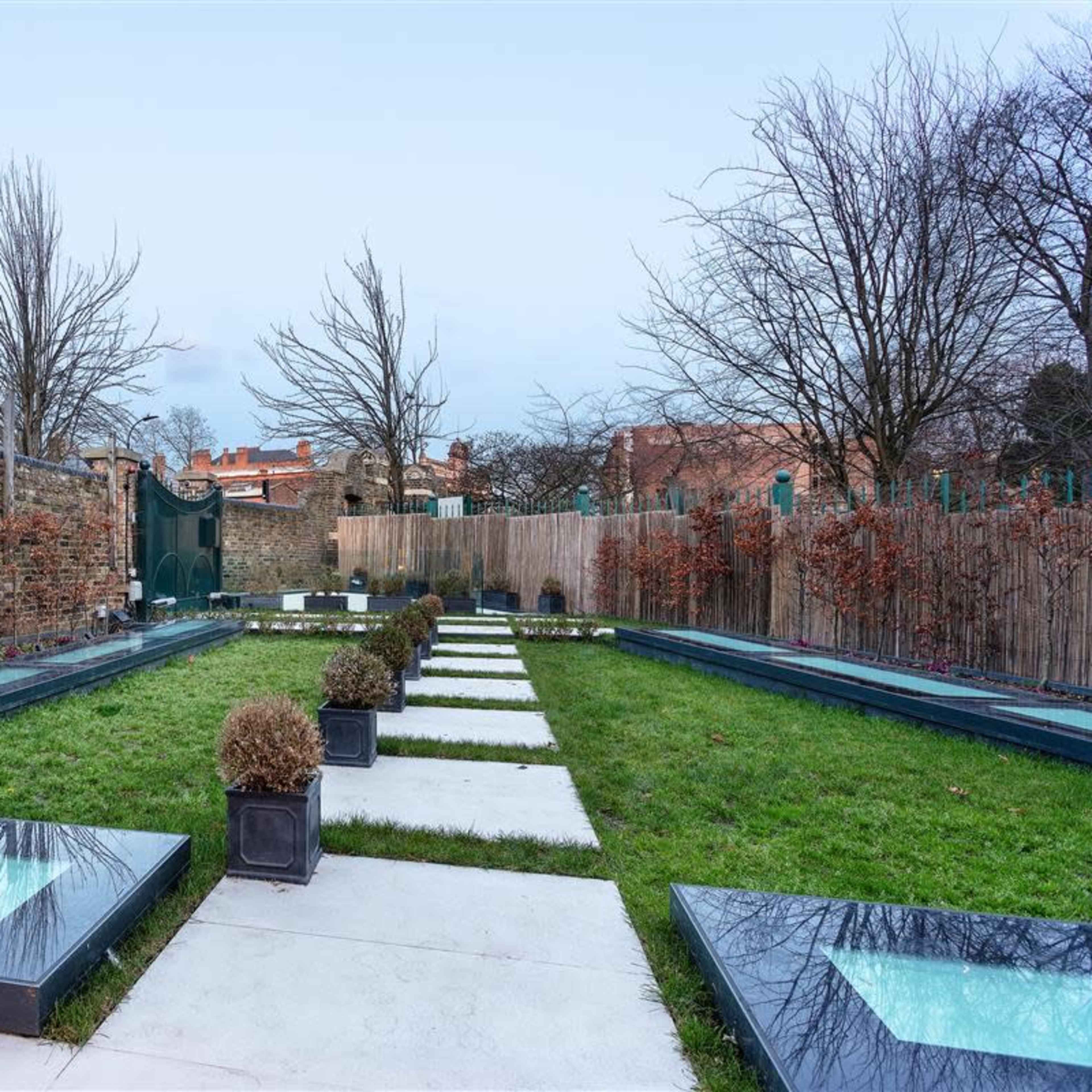 A landscaped garden featuring rectangular stepping stones, grassy areas, minimalist planters, and a stone wall backdrop.