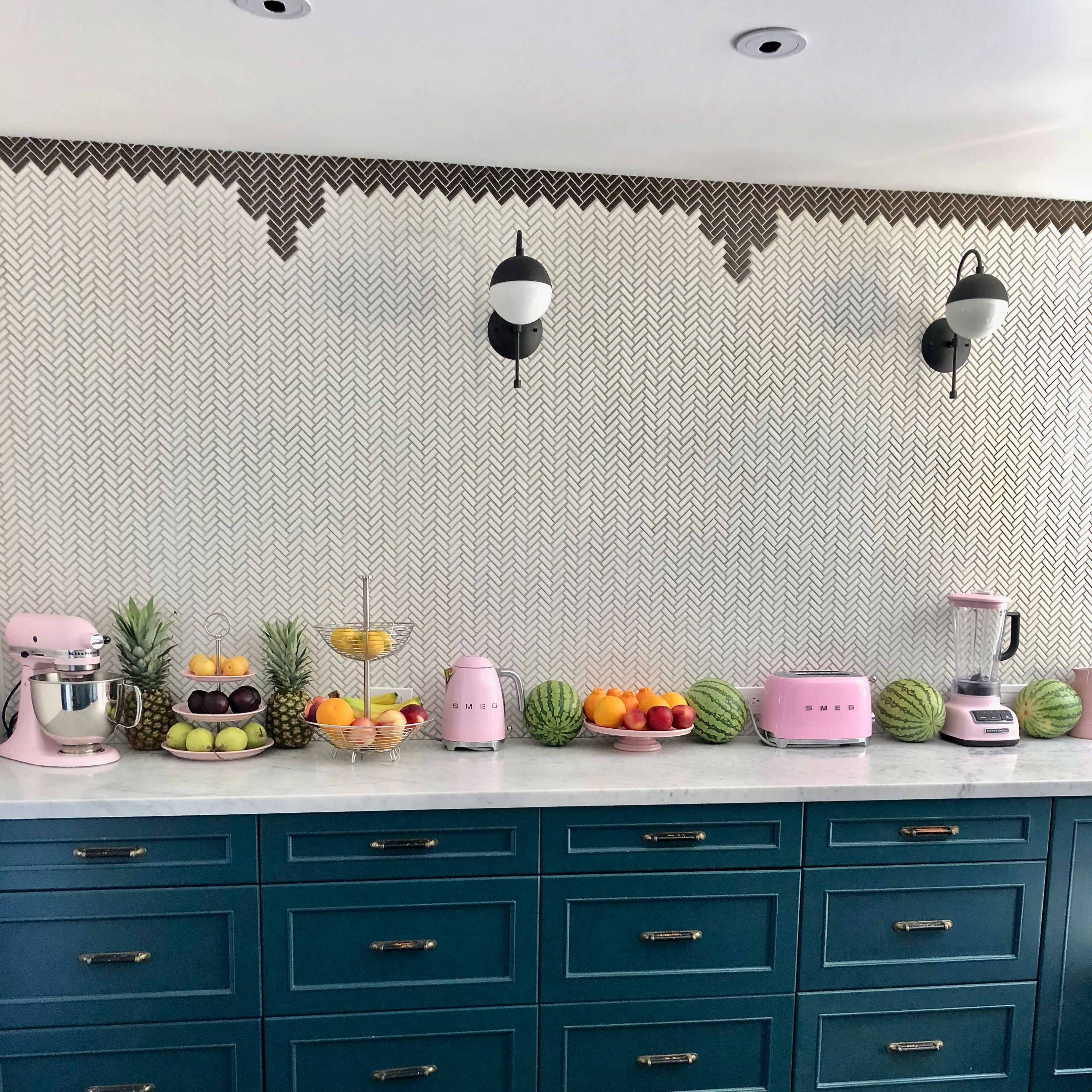 A modern kitchen features a teal cabinet with various fruits and pink appliances arranged on a marble countertop.