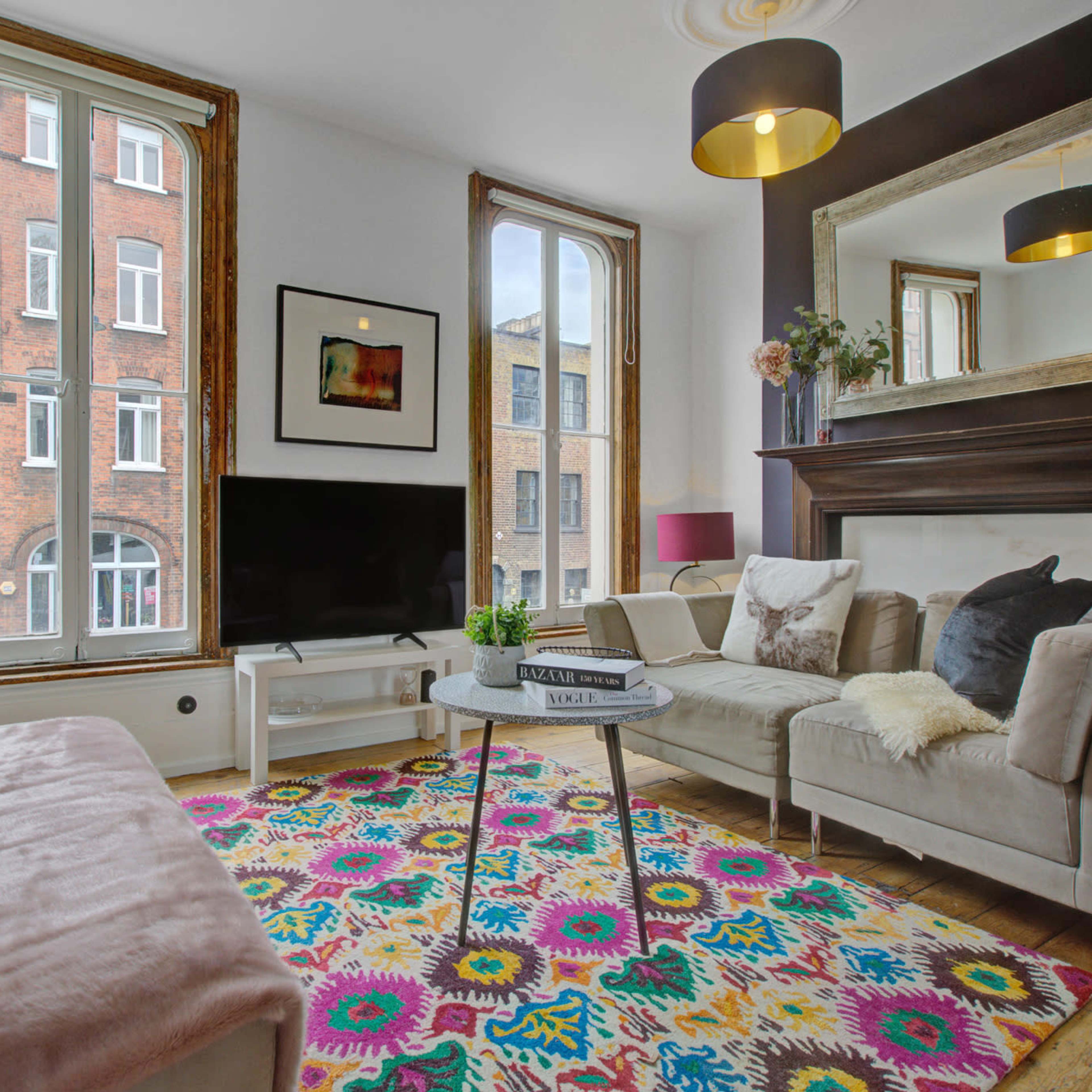 The living room features a light-colored sofa and armchair arranged around a small coffee table on a brightly patterned rug, with large windows providing views of a brick building outside.