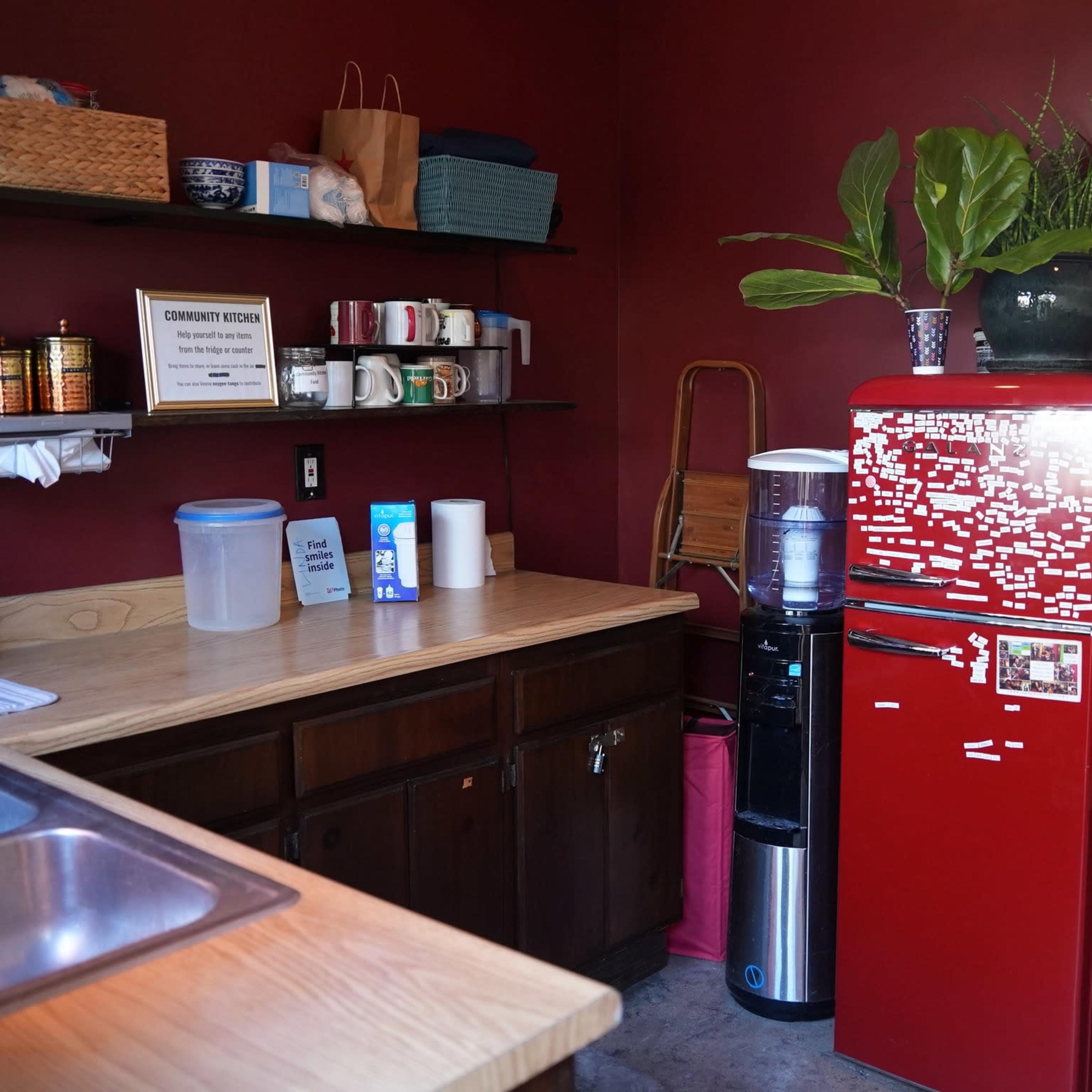 A small kitchen area features a red refrigerator, a water cooler, wooden shelves with various supplies, and a sink.