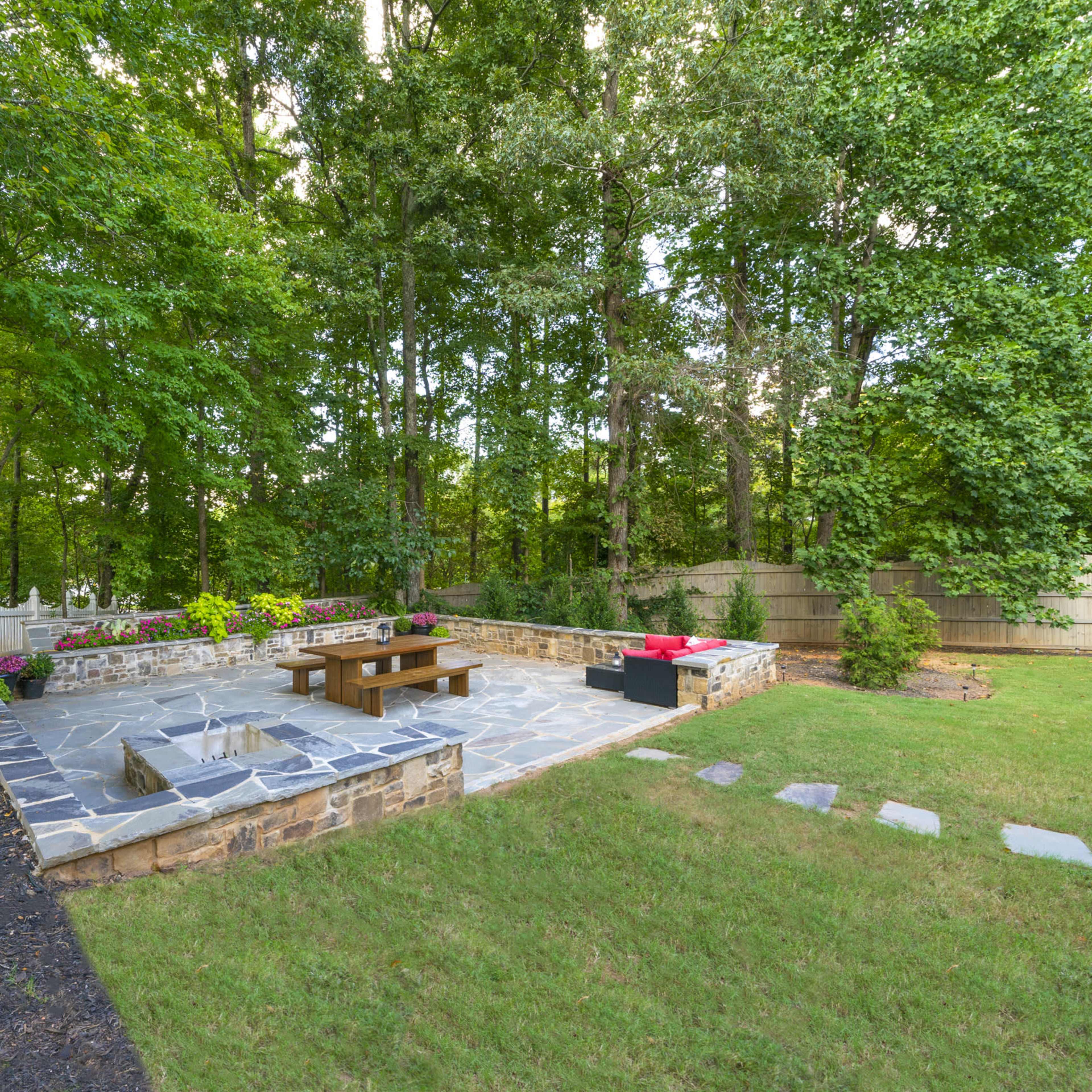 A stone patio with a wooden table and seating is surrounded by trees in a backyard.