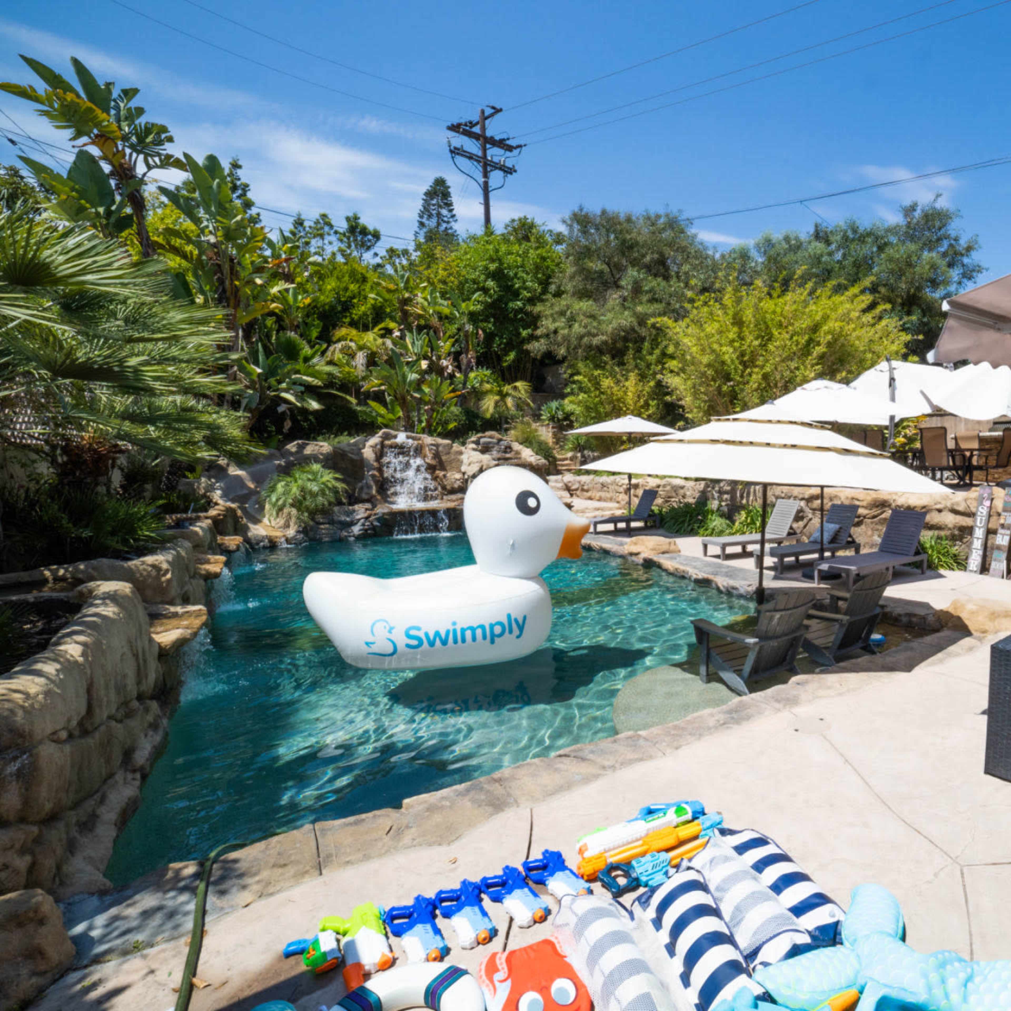 A large inflatable duck floats in a residential pool surrounded by lounge chairs and shaded seating areas.