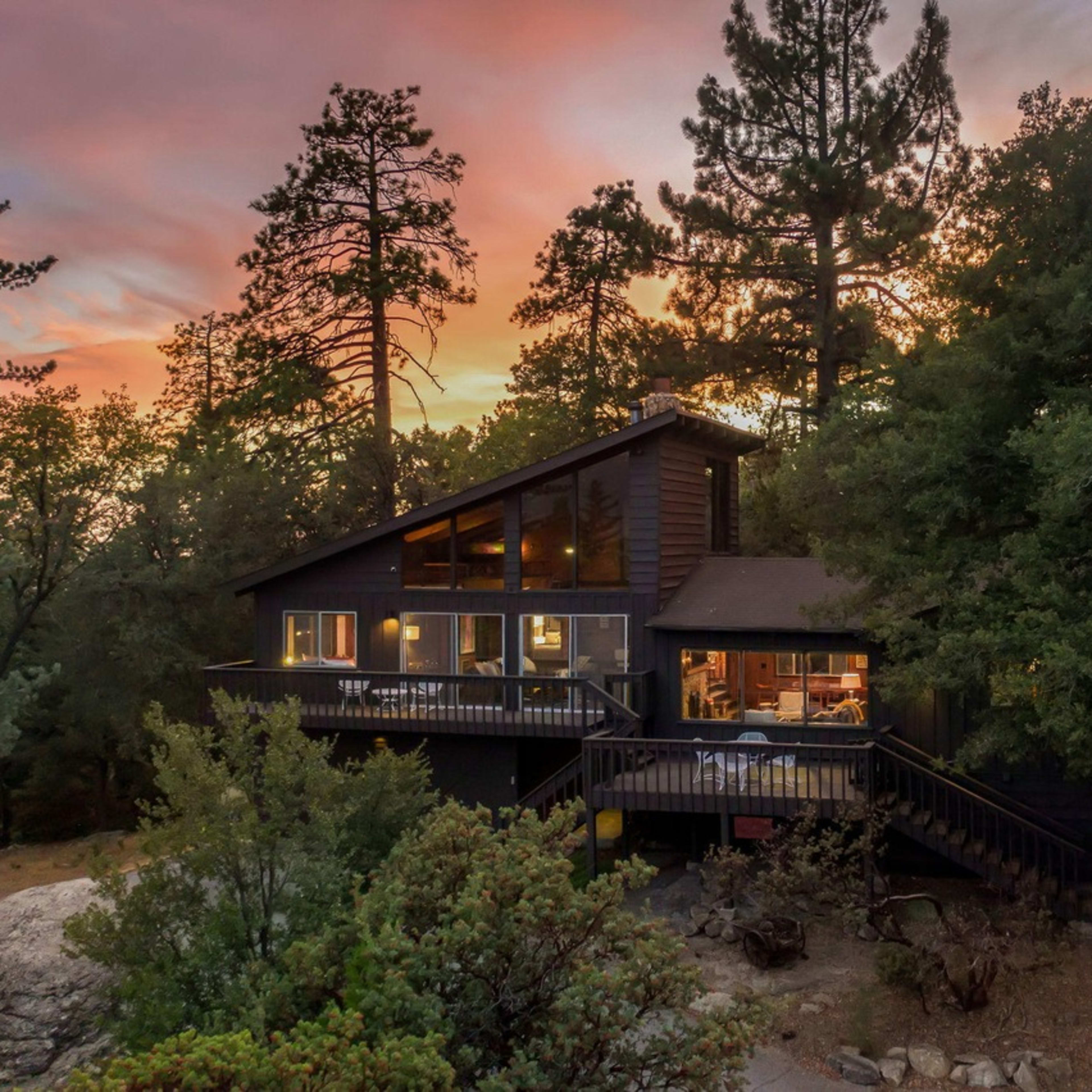 A modern cabin with large windows is surrounded by trees, set against a backdrop of a colorful sunset.