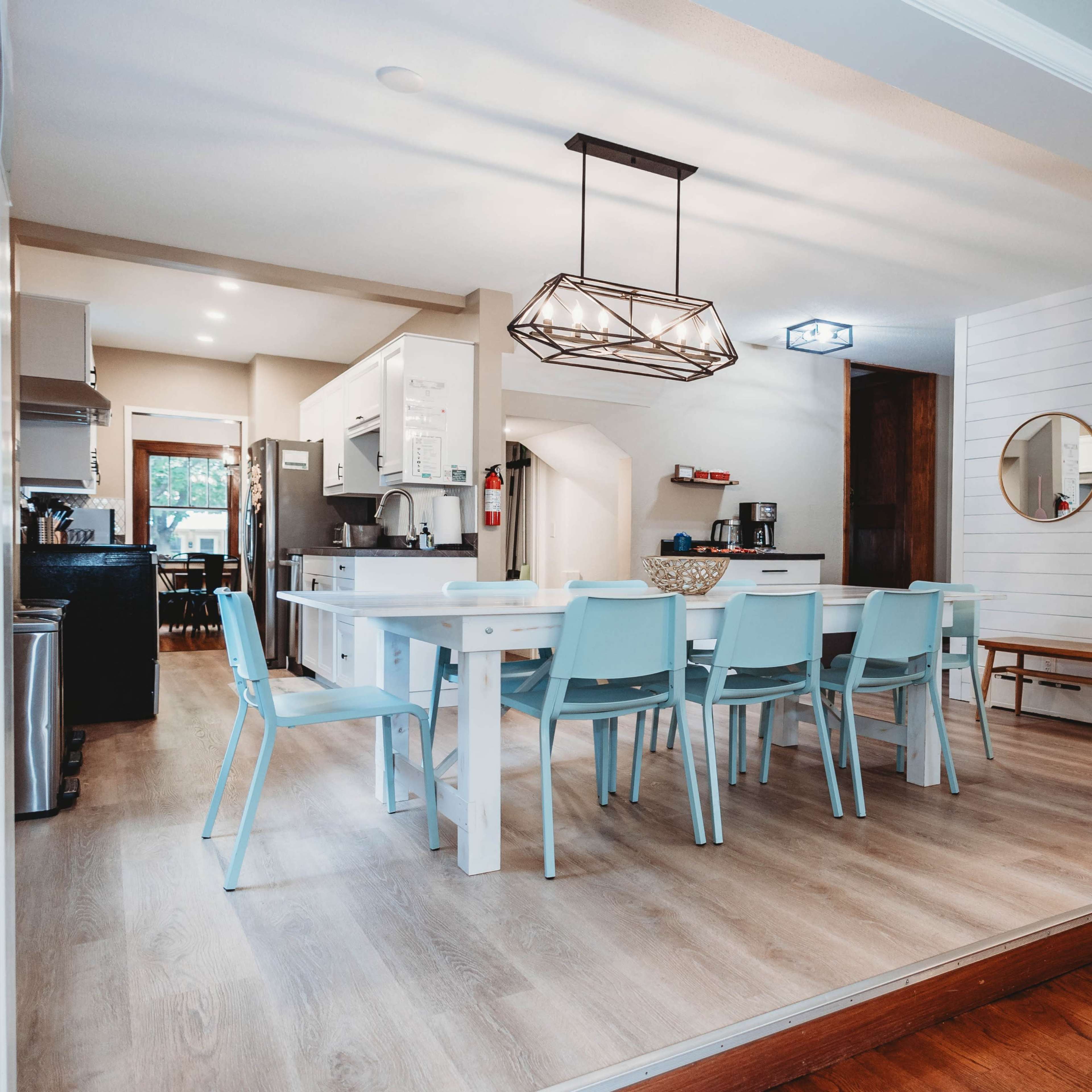 A modern kitchen and dining area with a long white table surrounded by light blue chairs, wooden flooring, and contemporary lighting fixtures.