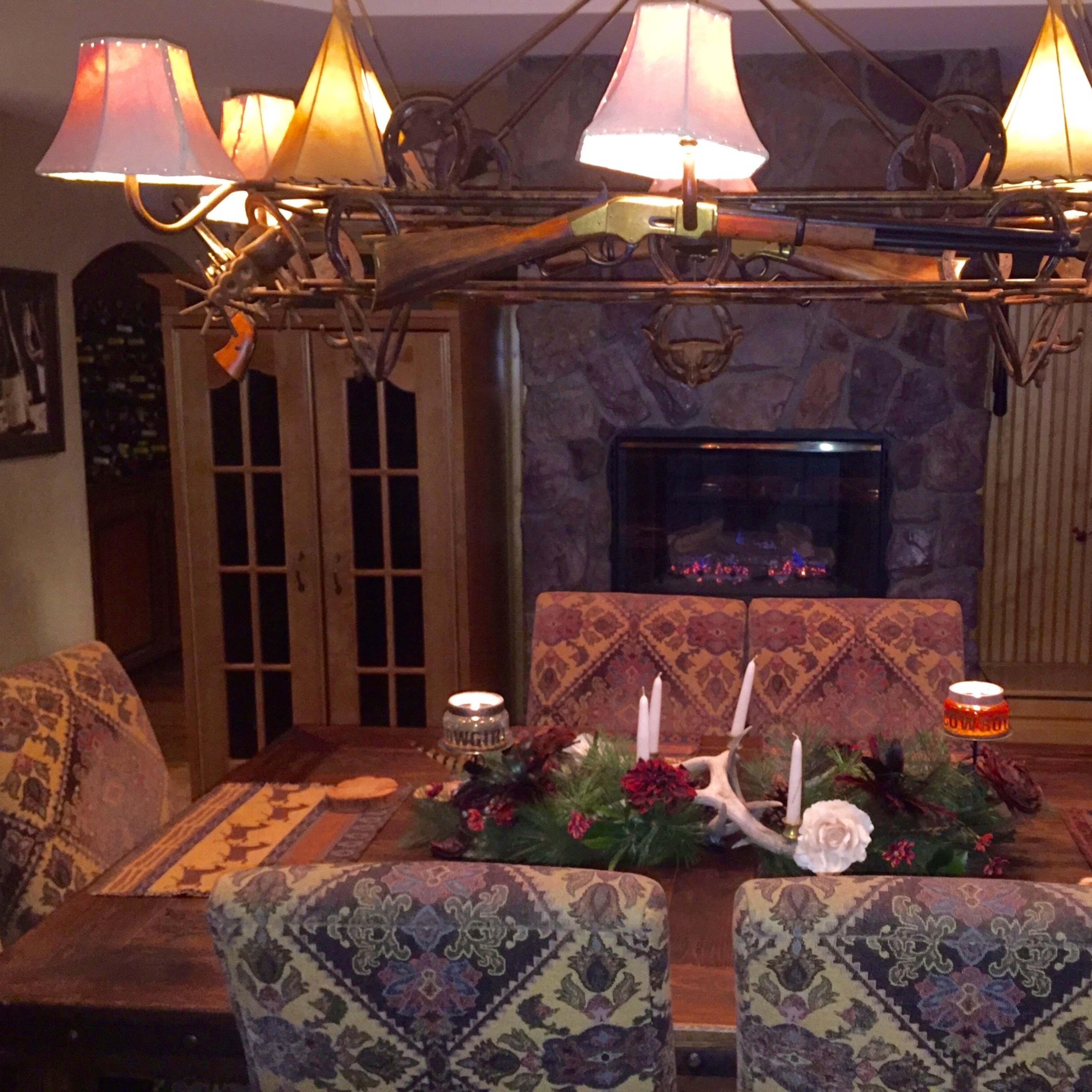 A rustic dining room features a large wooden table surrounded by patterned chairs, with a chandelier made of antlers overhead and a stone fireplace in the background.