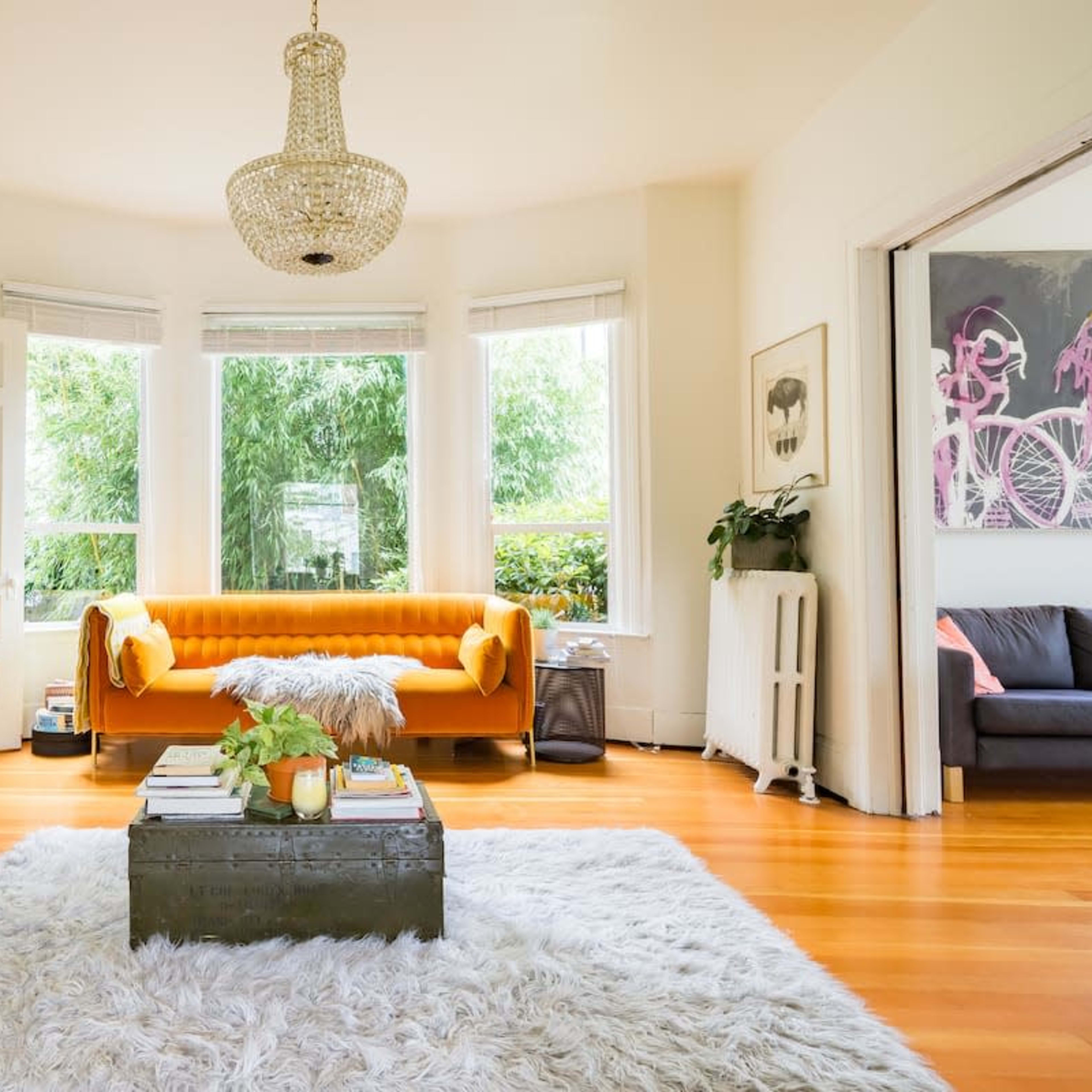 The scene depicts a bright living room featuring a yellow couch, a shaggy white rug, and large windows overlooking greenery.