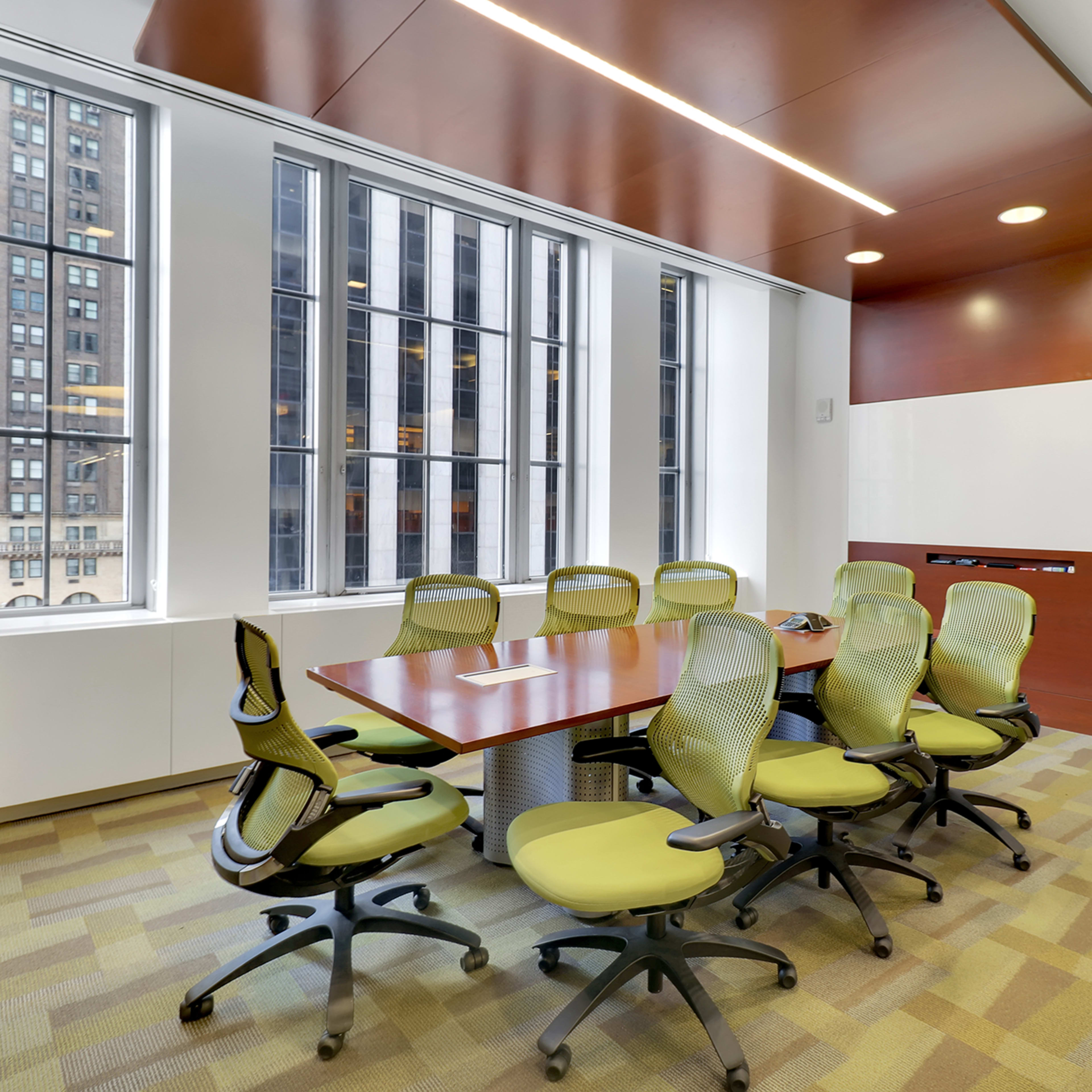 A modern conference room features a large wooden table surrounded by six green chairs, with large windows overlooking a cityscape.