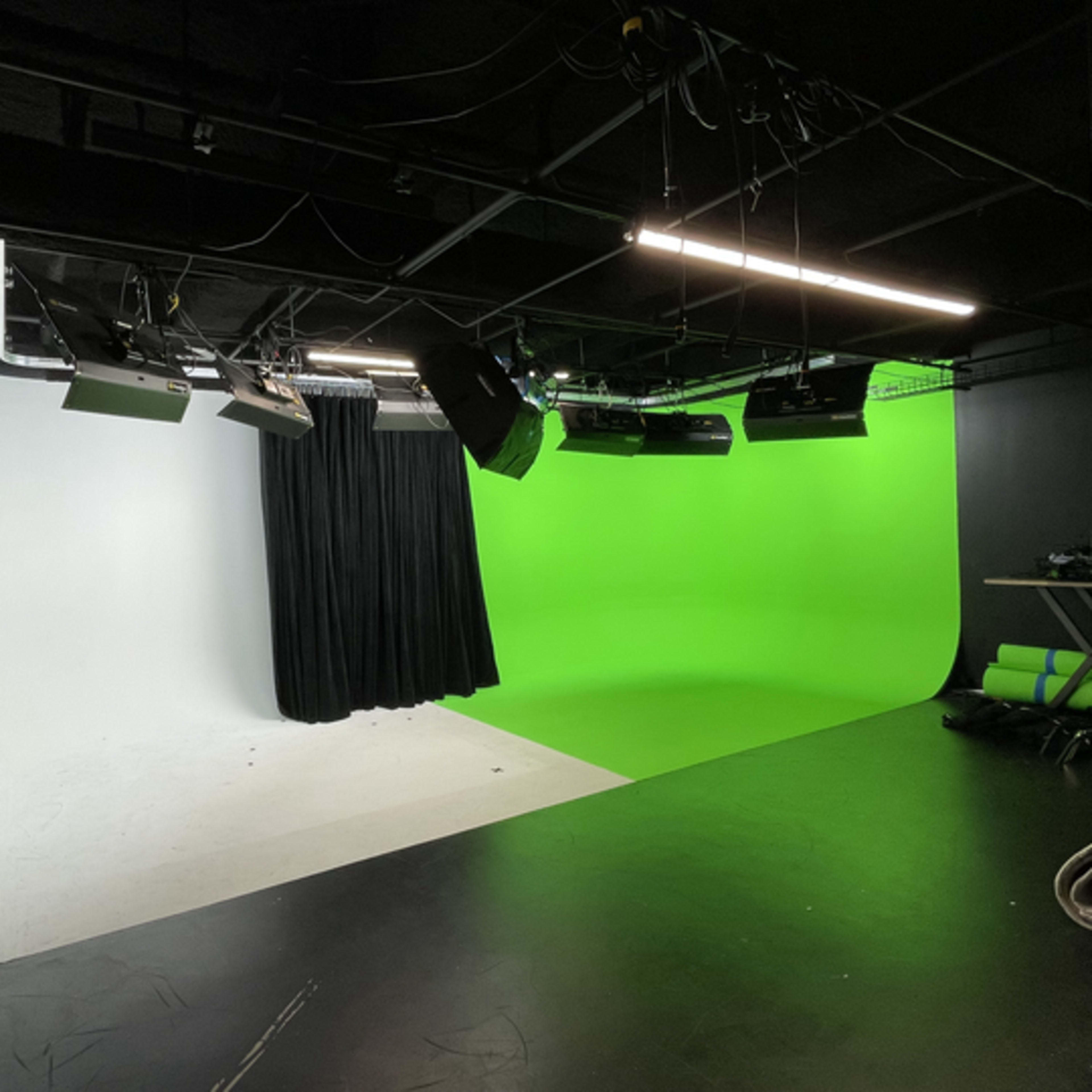 A studio with a split backdrop, featuring a white side and a green screen side, equipped with various lighting equipment and a black curtain.