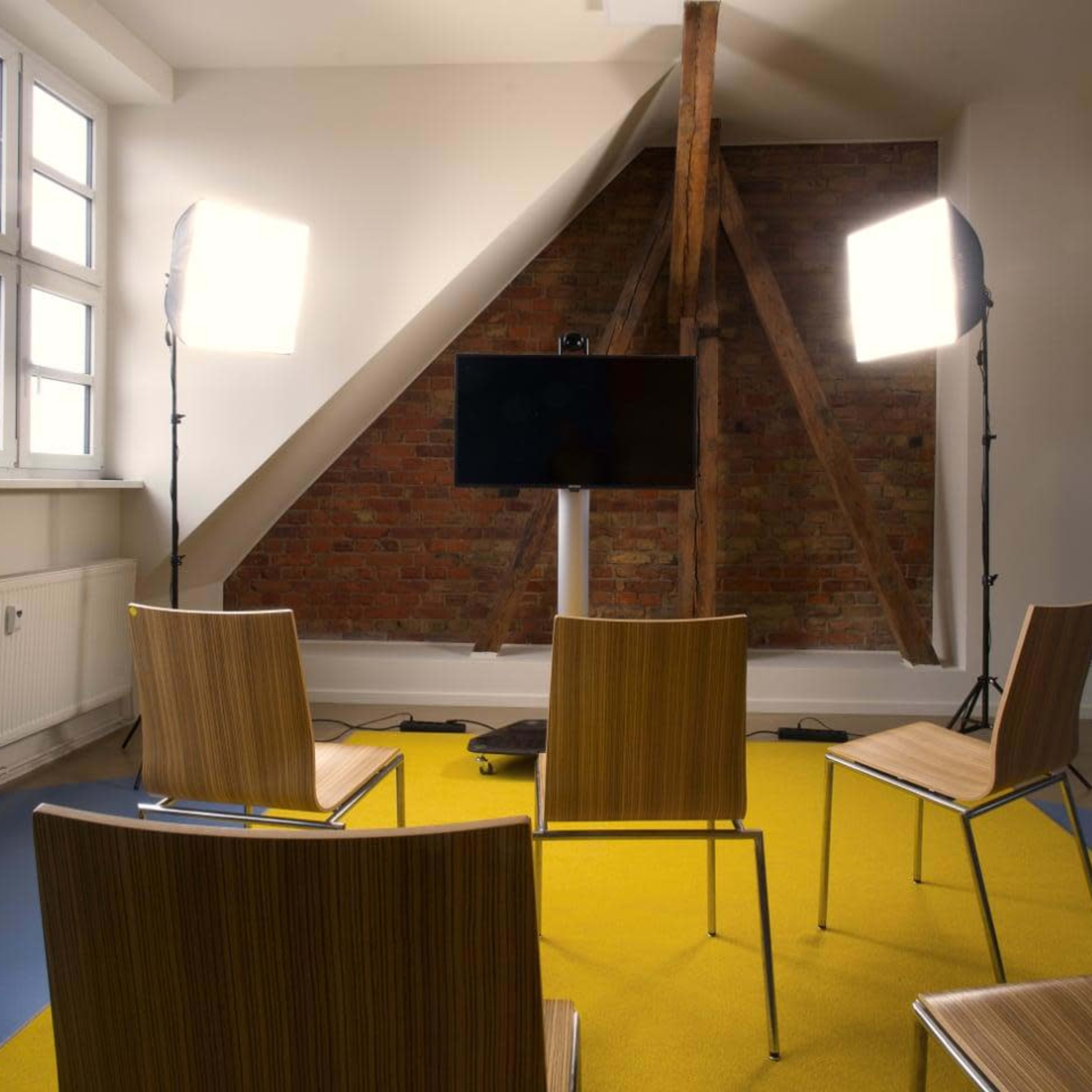 A meeting room is set up with four wooden chairs facing a television screen, illuminated by two bright lights, and featuring a yellow carpet with exposed brick walls.