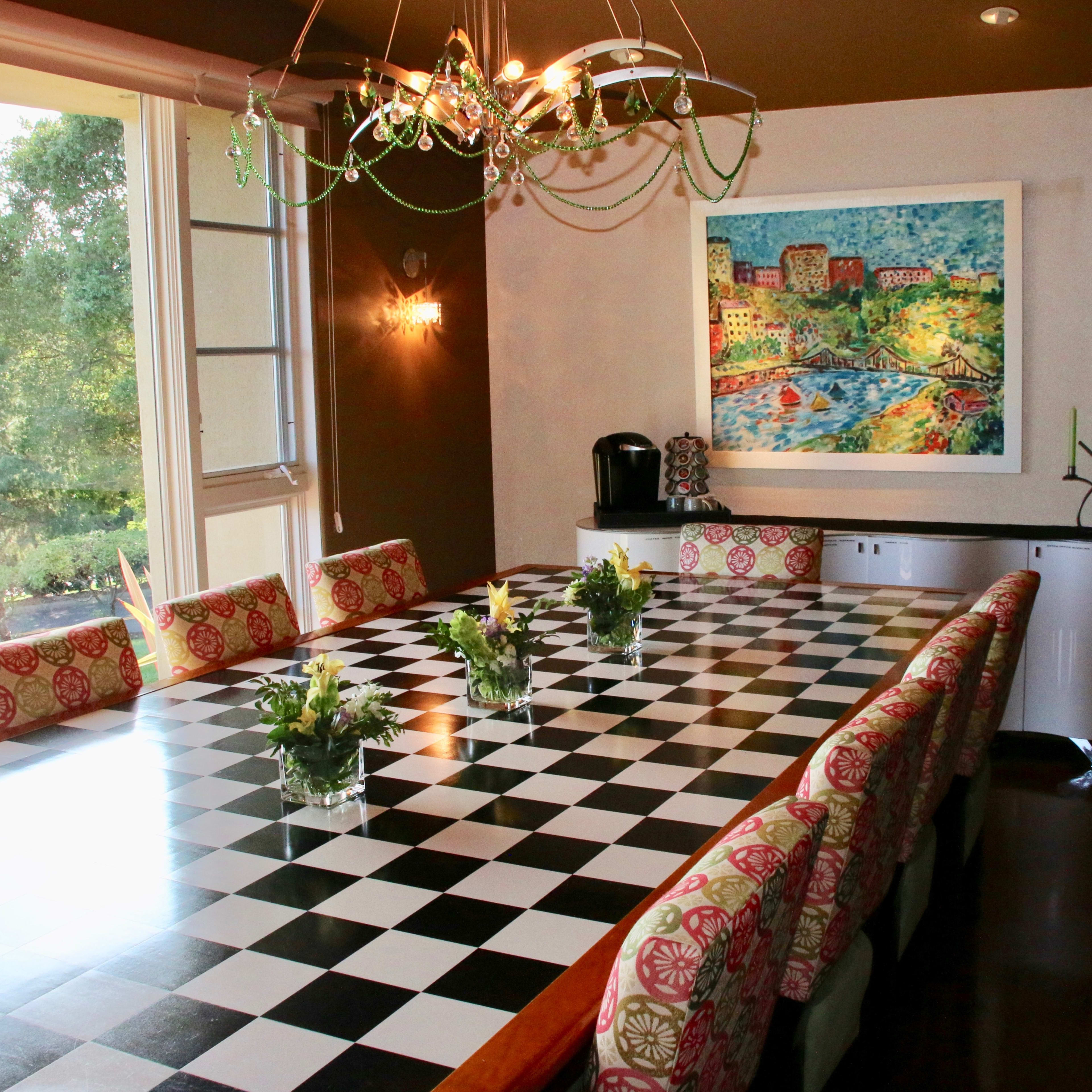 A large dining room features a checkered table with floral centerpieces, surrounded by patterned chairs, and a colorful artwork on the wall.