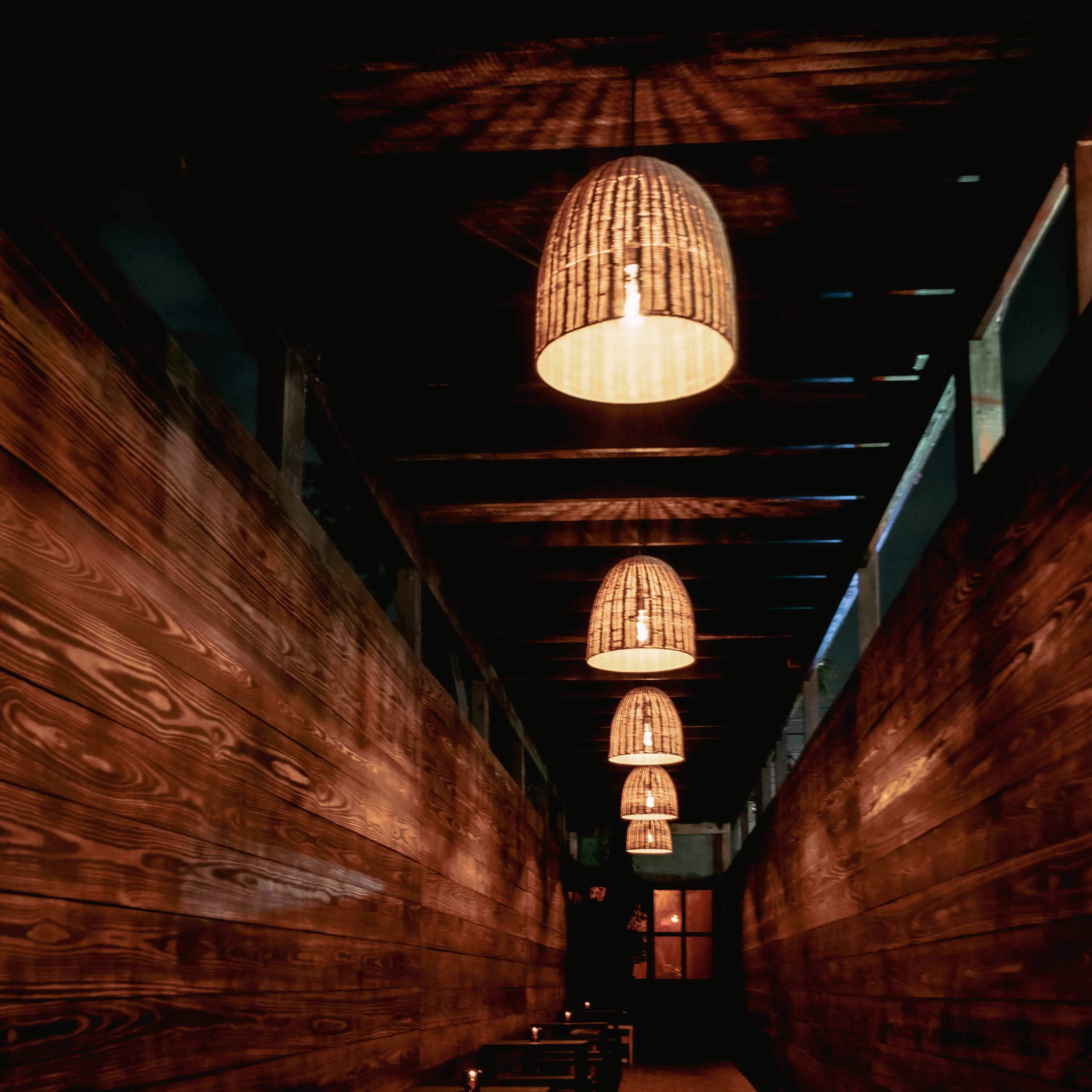 A dimly lit corridor features wooden walls with hanging woven light fixtures and small tables along the sides.