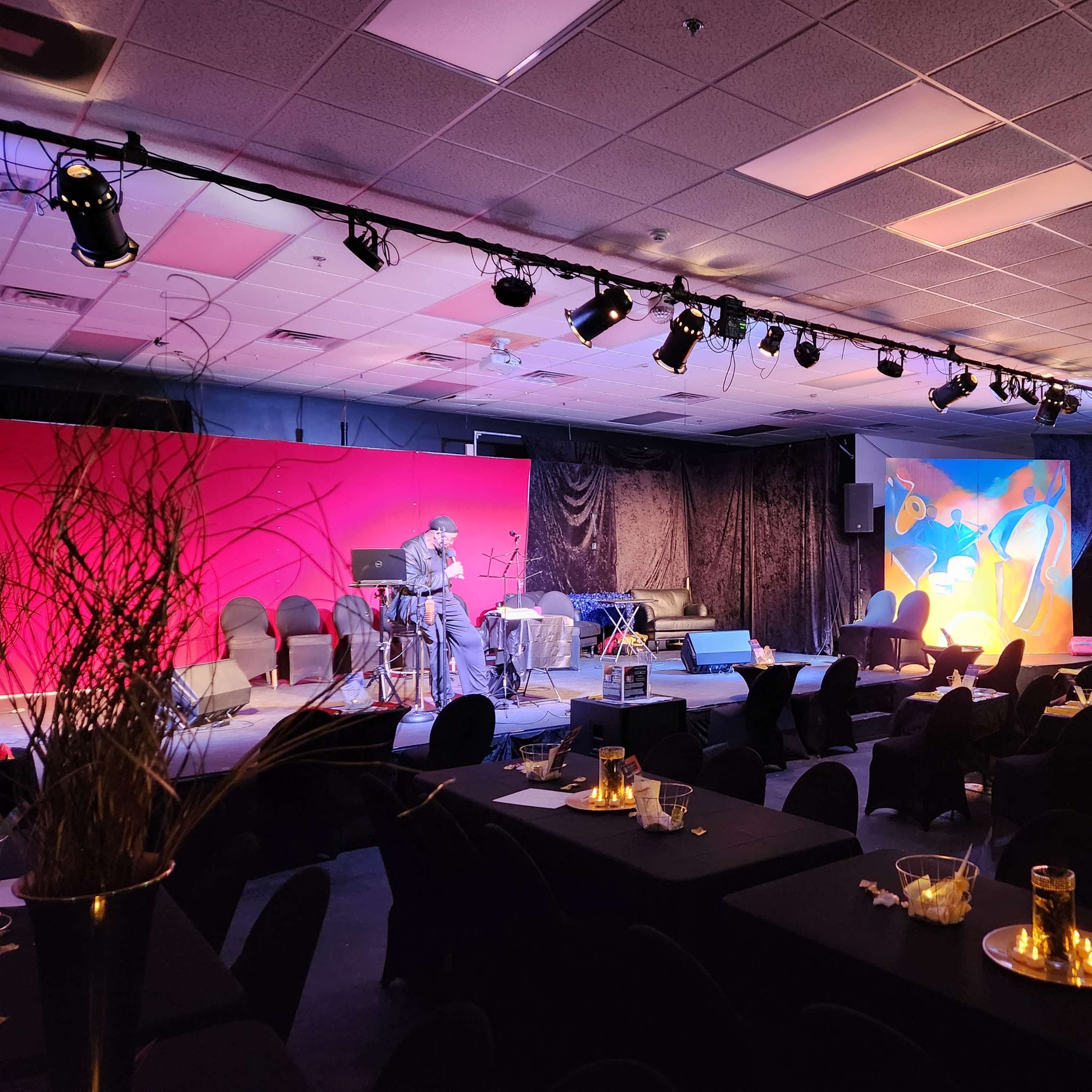 A stage with a musician performing is set up at the front of a dimly lit room filled with tables and chairs.